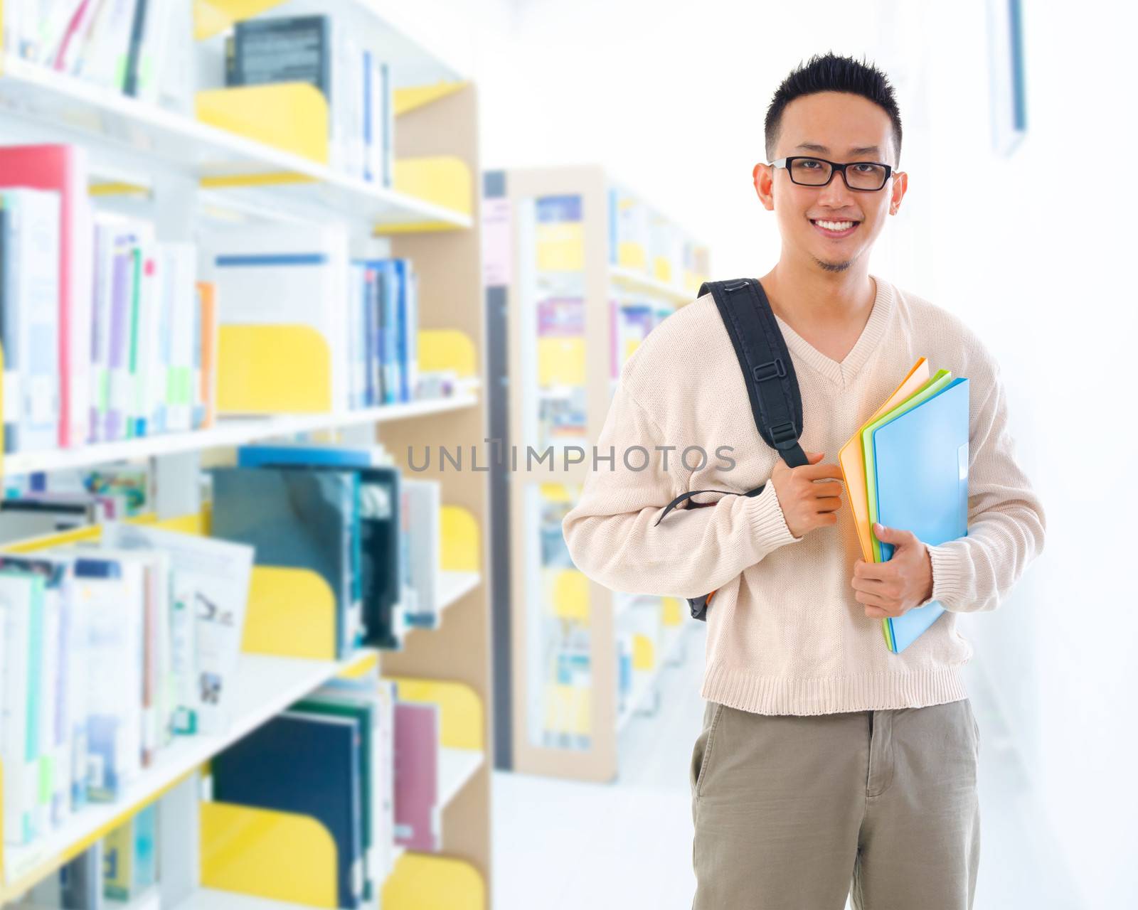 Good looking Southeast Asian adult student in casual wear with school bag carrying text books standing inside school library building.
