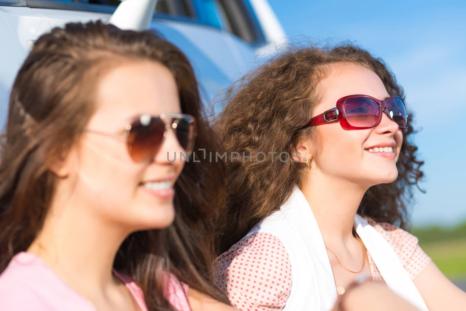 Two attractive young women wearing sunglasses, sitting next to the car