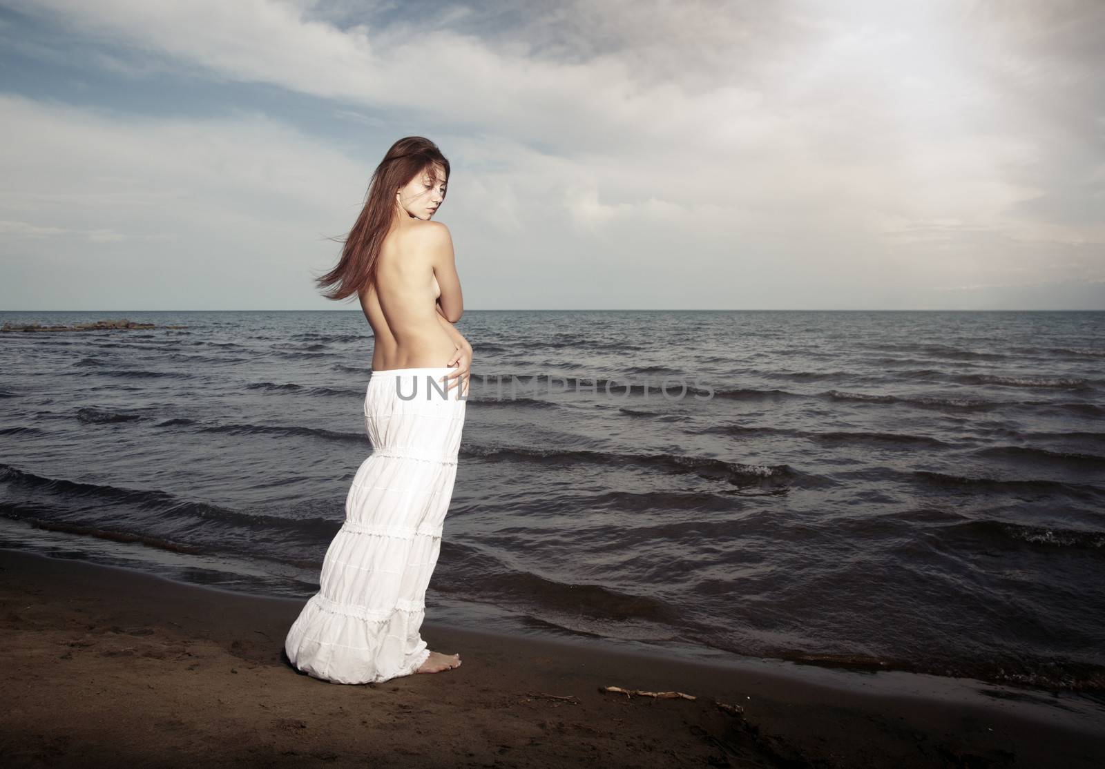 Topless lady with long hairs standing near the cold sea. Horizontal photo