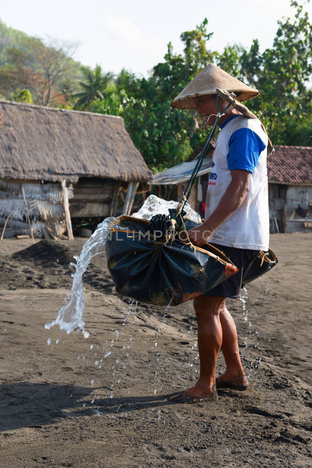 BALI, INDONESIA - SEP 26: Manual male worker spreads sea water on black volcanic sand for salt production on Sep 26, 2012 in Amuk Bay, Bali, Indonesia. It is a unique tradition dating back over 900 years. 