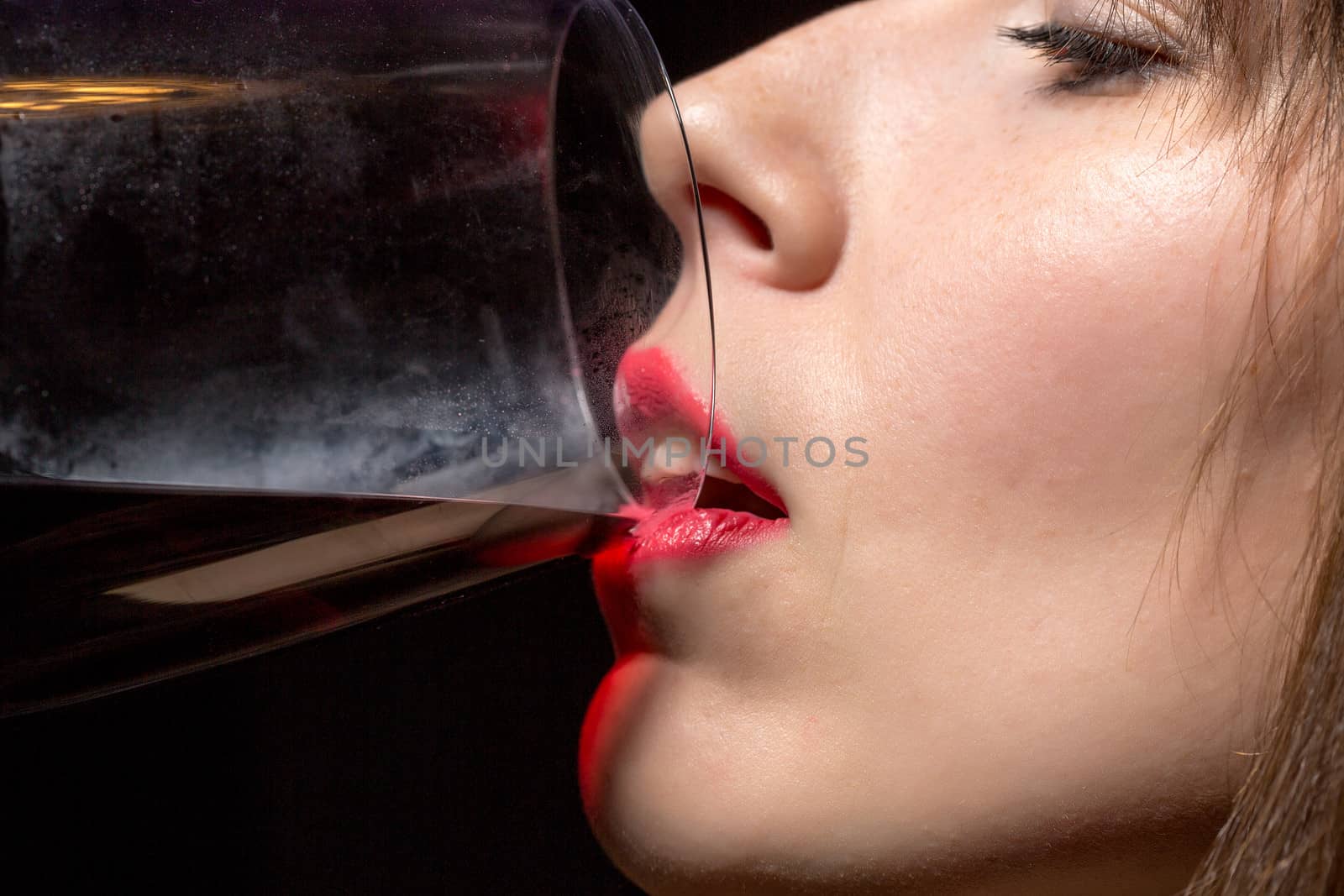 Closeup of the mouth and sexy red lips of a young woman drinking red wine from a wineglass against a dark background