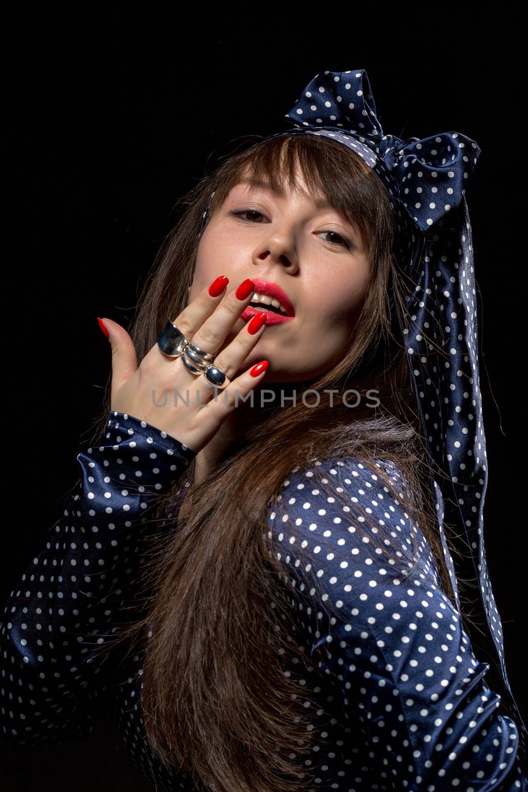 Elegant sensual woman with long brunette hair wearing stylish polka dot evening dress applying red lipstick to her lips against a dark background