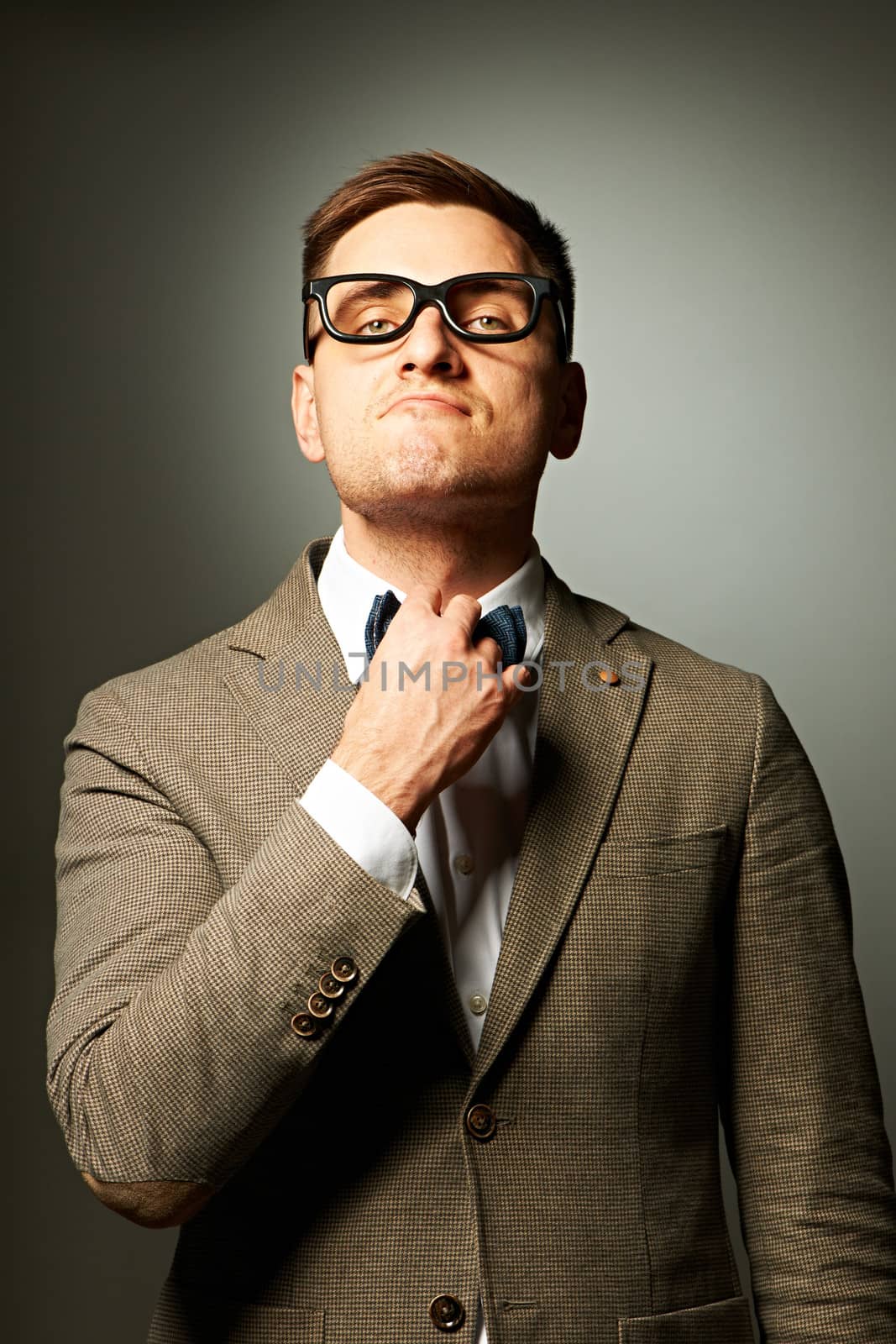 Confident nerd in eyeglasses adjusting his bow-tie against grey background