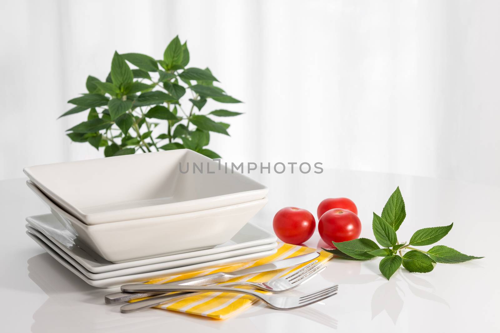 Plates, kitchen utensils, herbs and tomatoes on a white table.