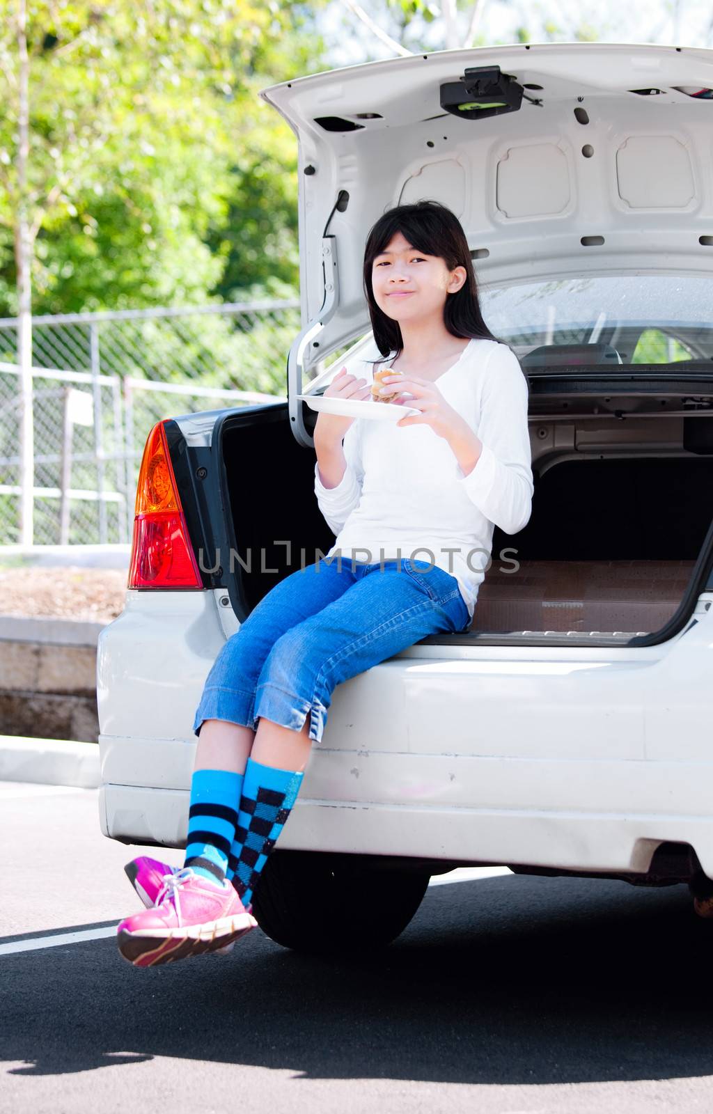 Preteen biracial girl sitting in opened car trunk, eating lunch