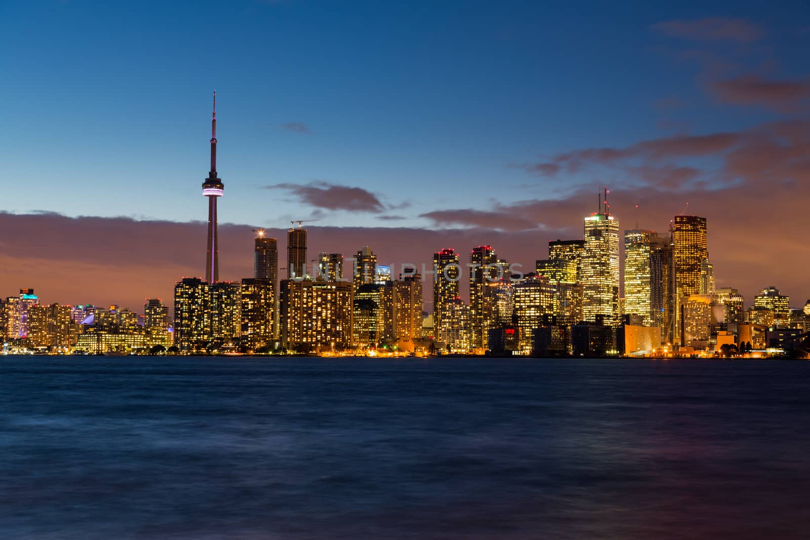 Light remains in sky with low orange clouds over skyline of Toronto with Lake Ontario in foreground and skyscrapers of city illuminated