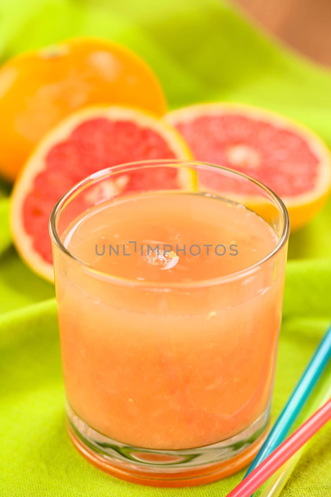 Freshly squeezed juice of the pink-fleshed grapefruit with colorful drinking straws on the side and grapefruits in the back on green fabric (Selective Focus, Focus on the bubble on the middle of the juice)