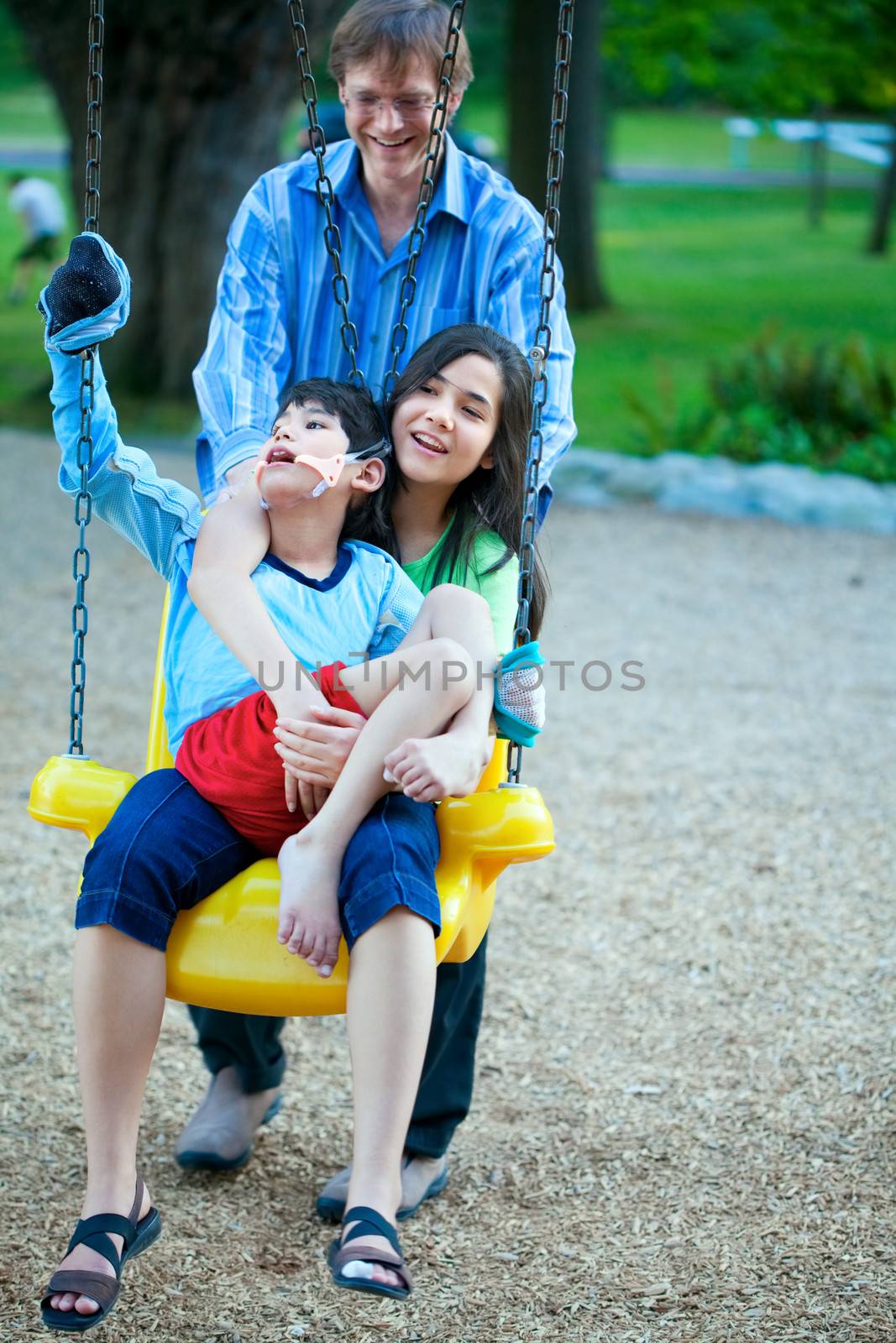 Big sister holding disabled brother on special needs swing at playground in park as father pushes. Child has cerebral palsy.