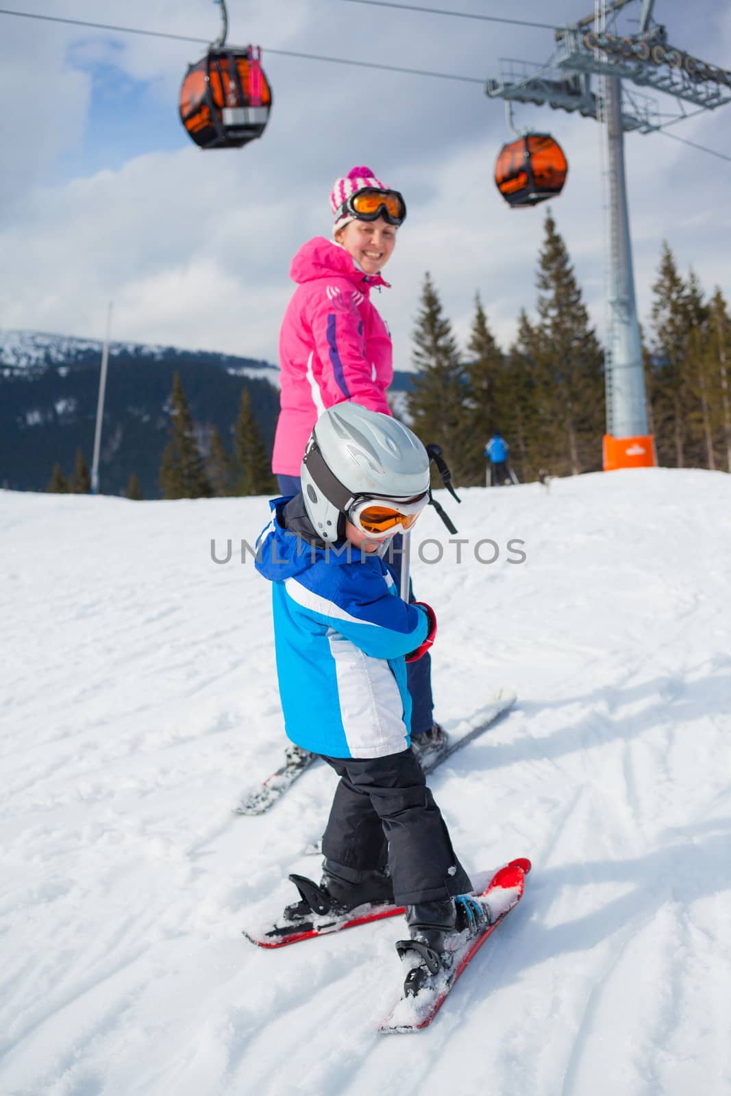 Happy little boy studying to ski in ski goggles and a helmet with his mother
