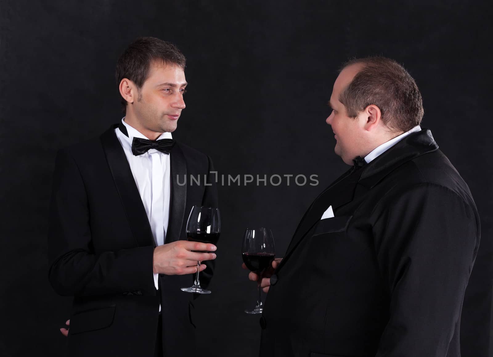 Two stylish businessman in tuxedos with glasses of red wine by Discovod