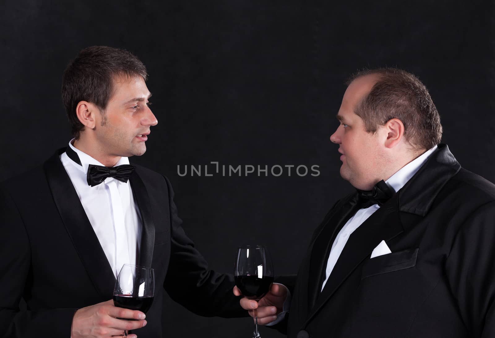 Two stylish businessman in tuxedos with glasses of red wine, on black background