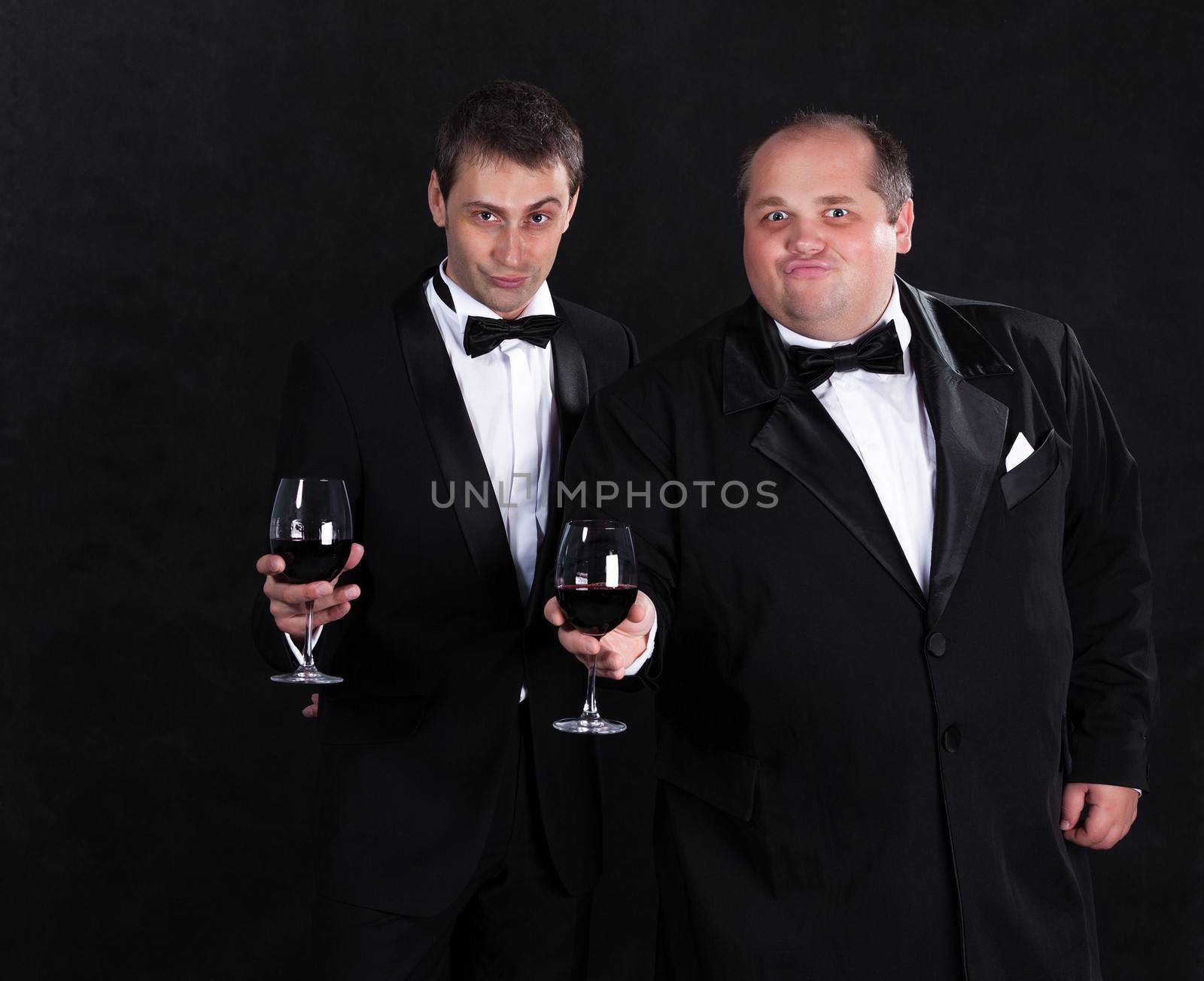 Two stylish businessman in tuxedos with glasses of red wine, on black background