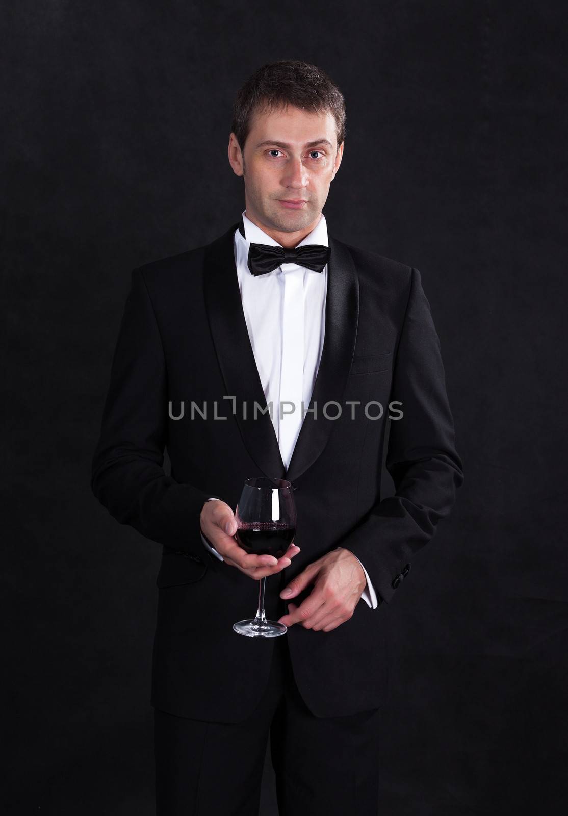 Stylish man in elegant black tuxedo with glass red wine, on black background