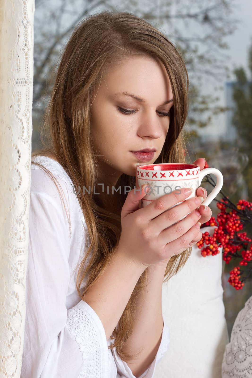 Portrait of charming girl in sweater holding cup and looking through window