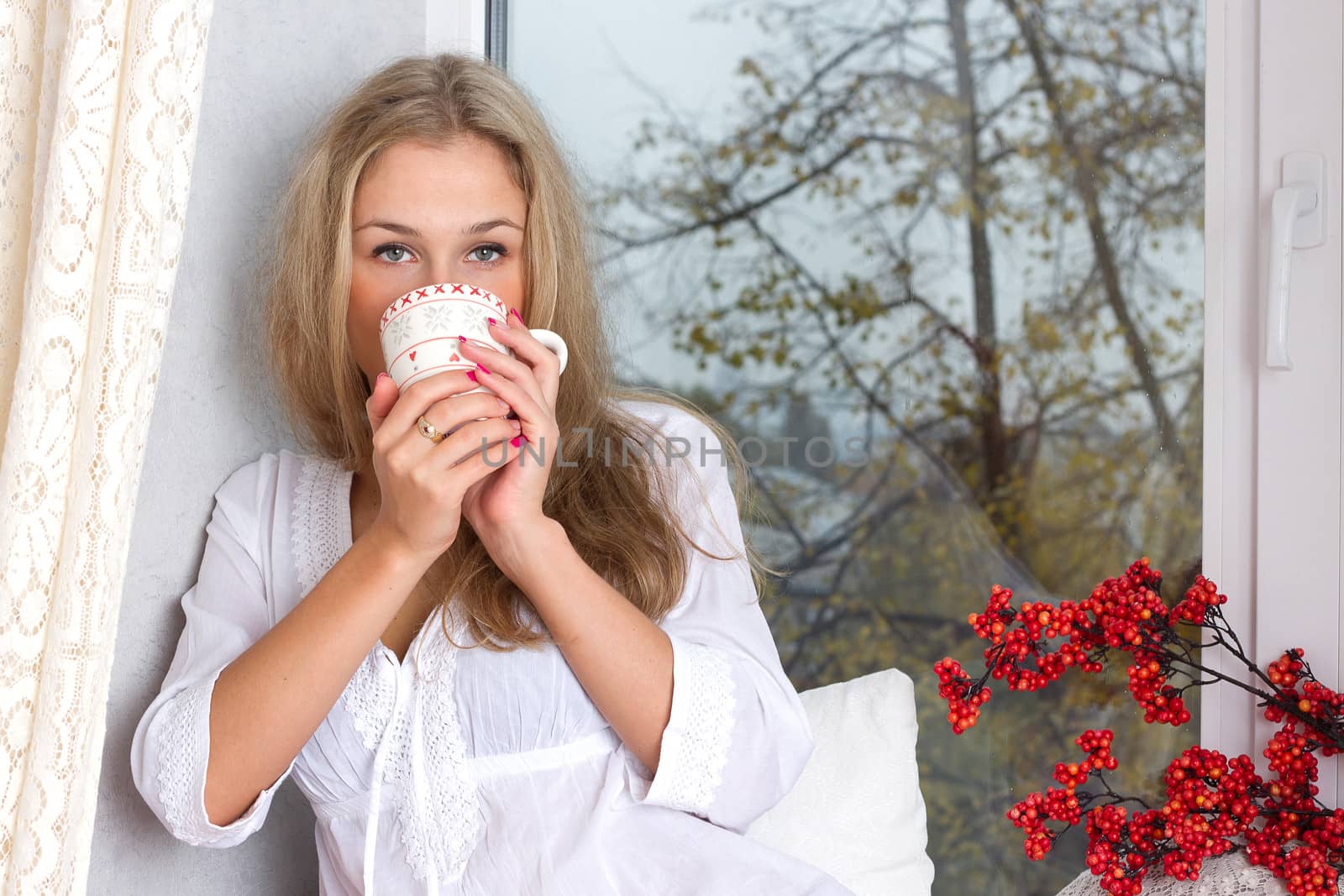 Portrait of charming girl in sweater holding cup and looking through window