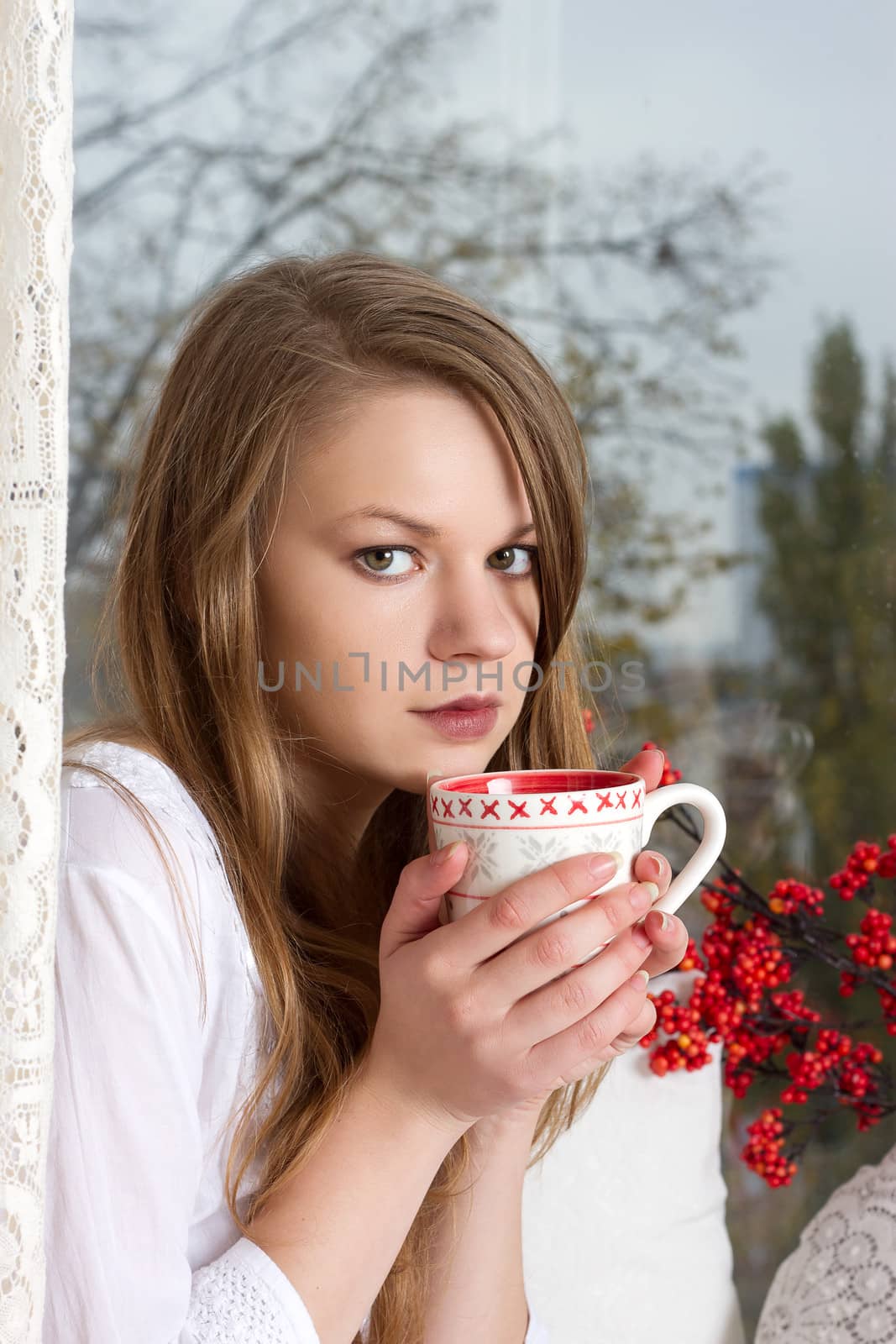 Portrait of charming girl in sweater holding cup and looking through window
