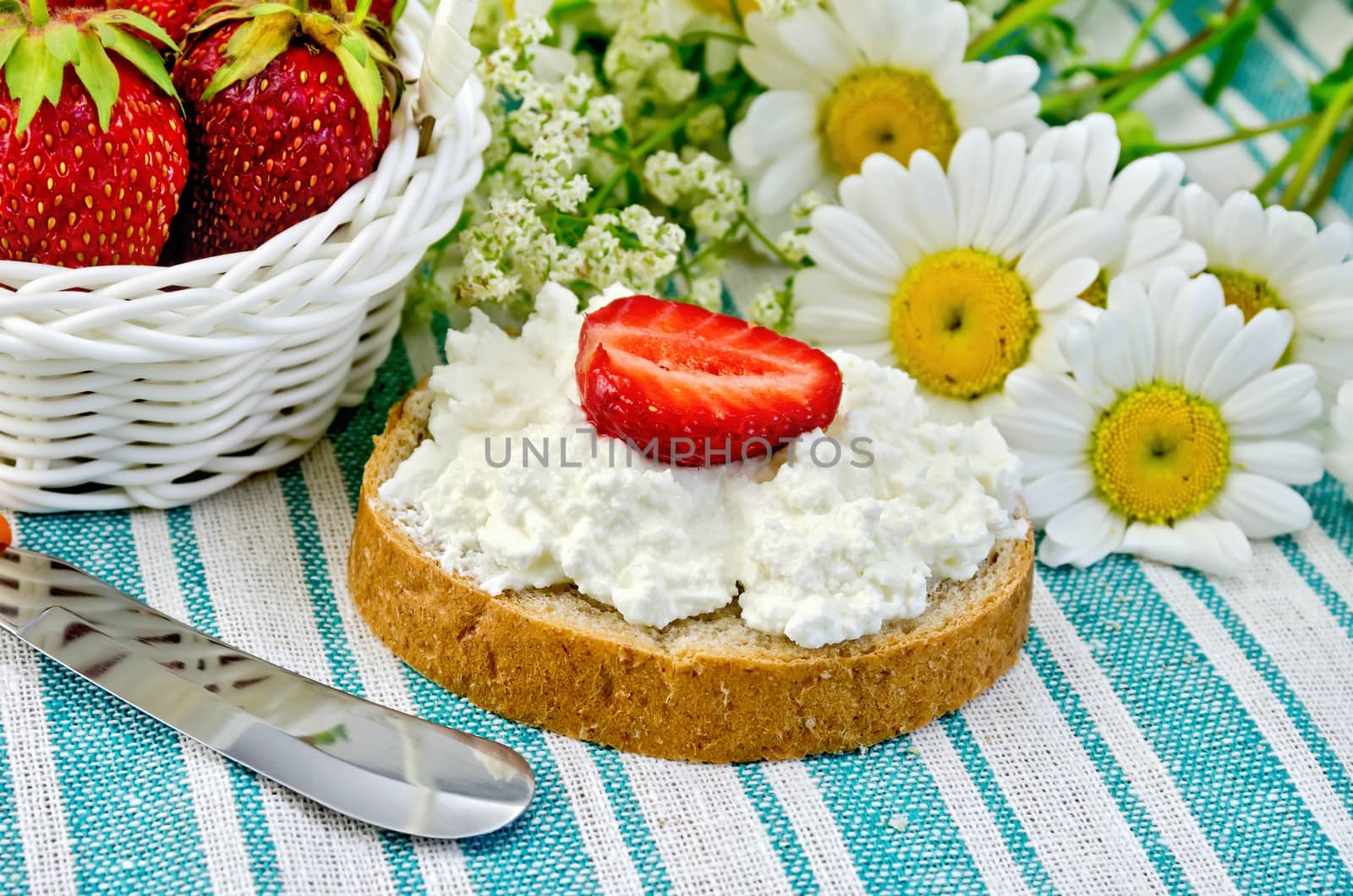 Hunk of bread with cottage cheese cream and strawberries, a basket with berries, a knife, a bouquet of daisies on a green striped cloth