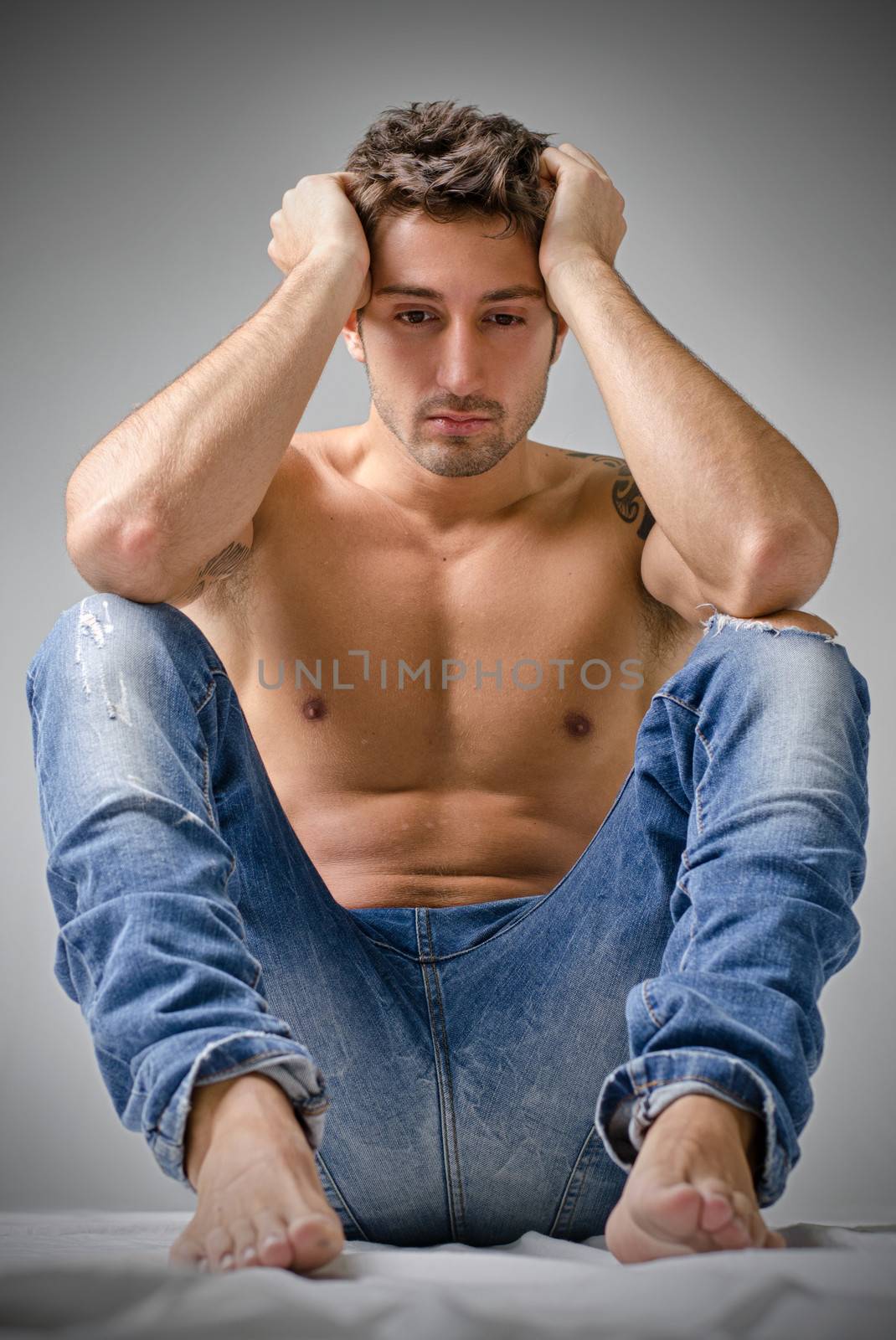 Depressed or sad handsome young man shirtless in jeans, sitting on floor with head in his hands