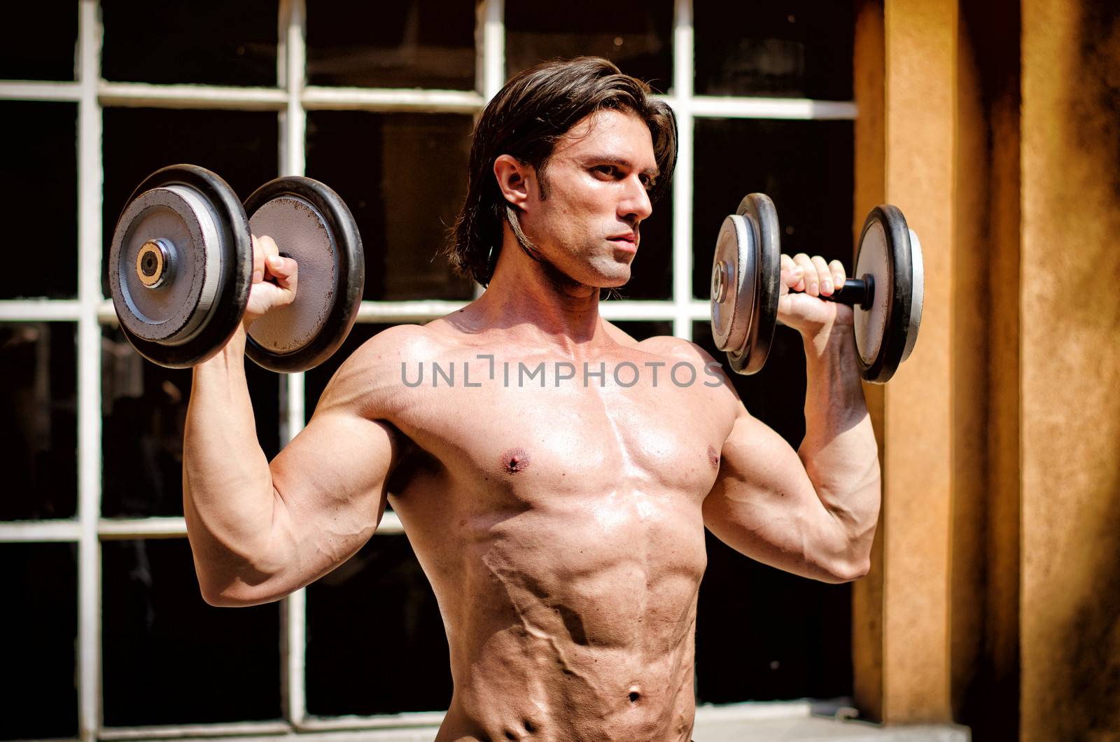 Muscular young bodybuilder shirtless outdoors, exercising biceps with dumbbells