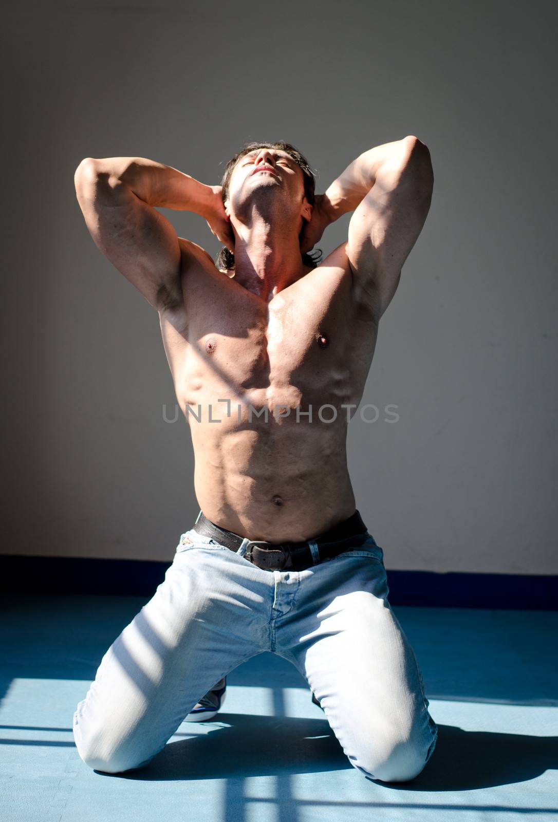 Attractive muscleman kneeling shirtless on grey background with hands behind head, looking up