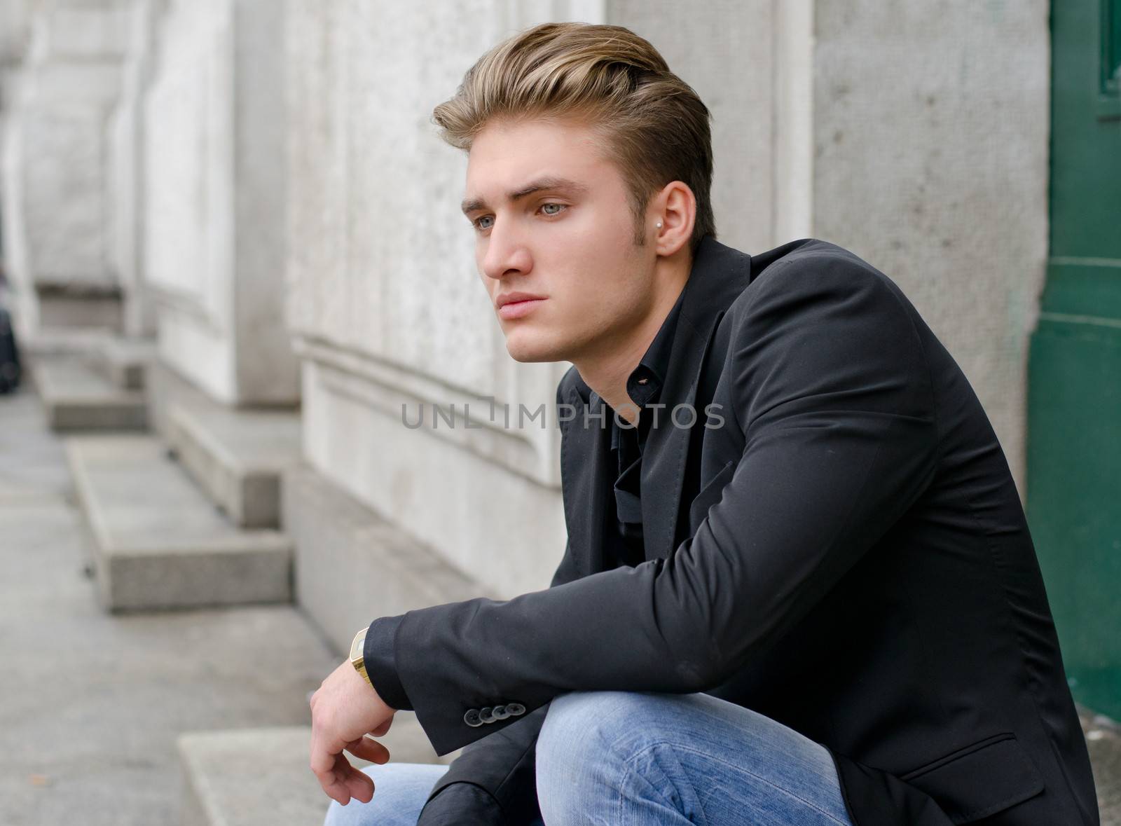 Attractive blond young man in jeans and jacket, sitting outdoors looking off camera