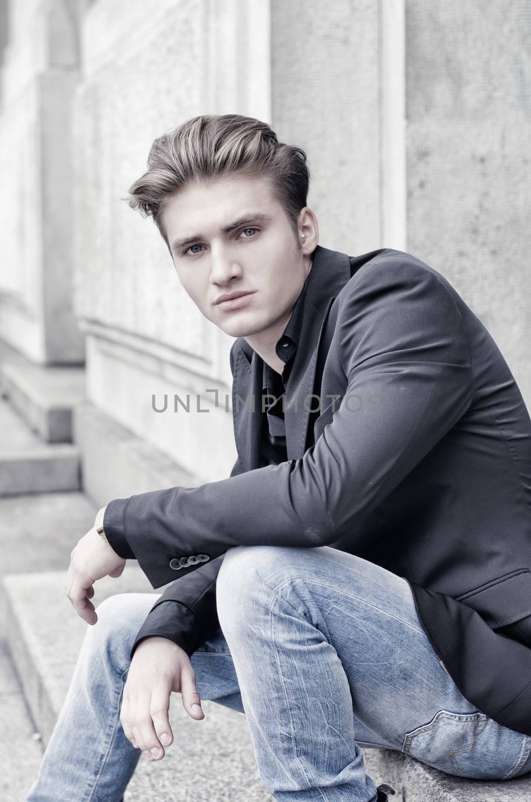 Attractive blond young man in jeans and jacket, sitting outdoors looking in camera