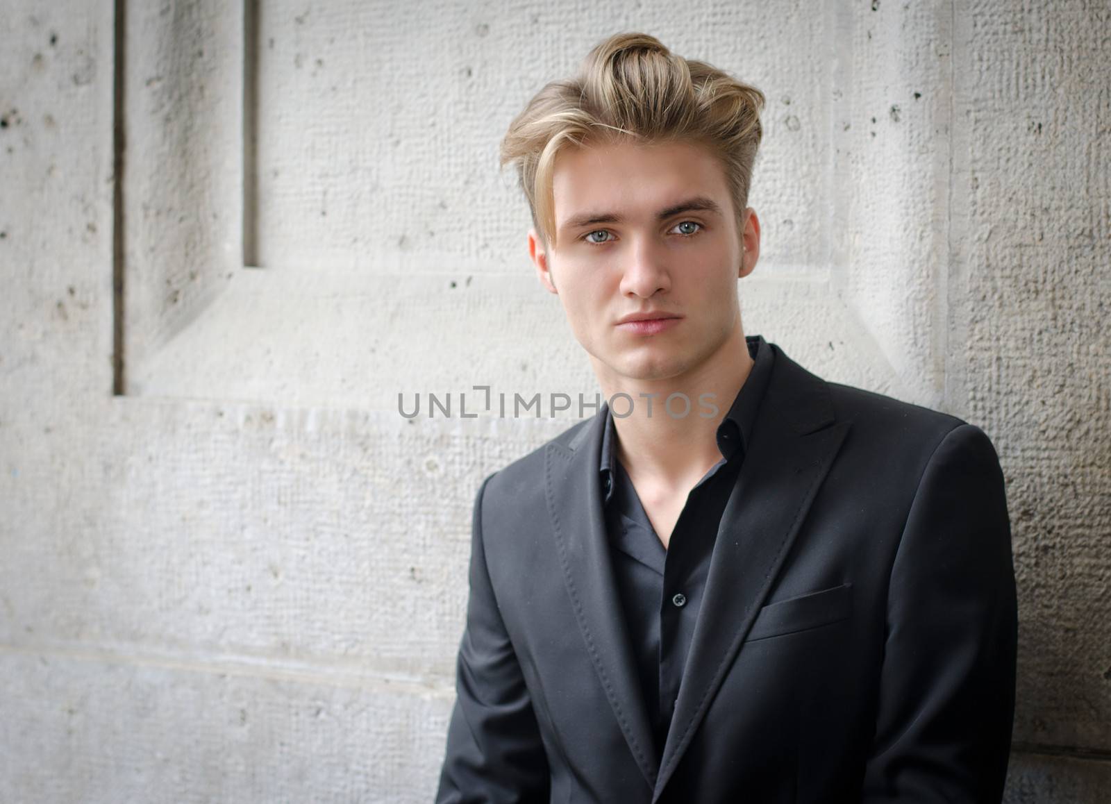 Attractive blond young man in shirt and jacket, standing against wall
