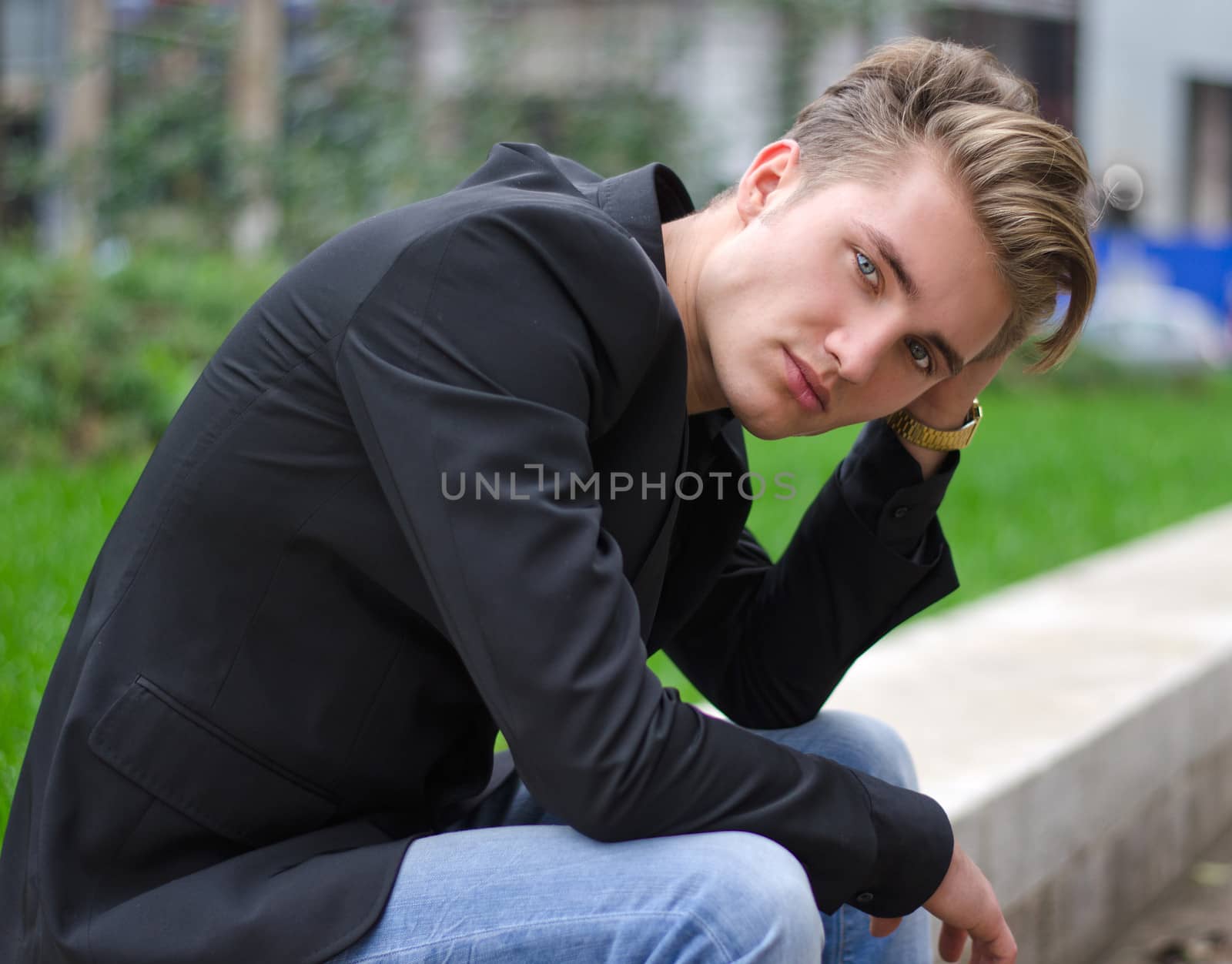 Serious or sad blond young man in jeans and jacket, sitting outdoors looking in camera