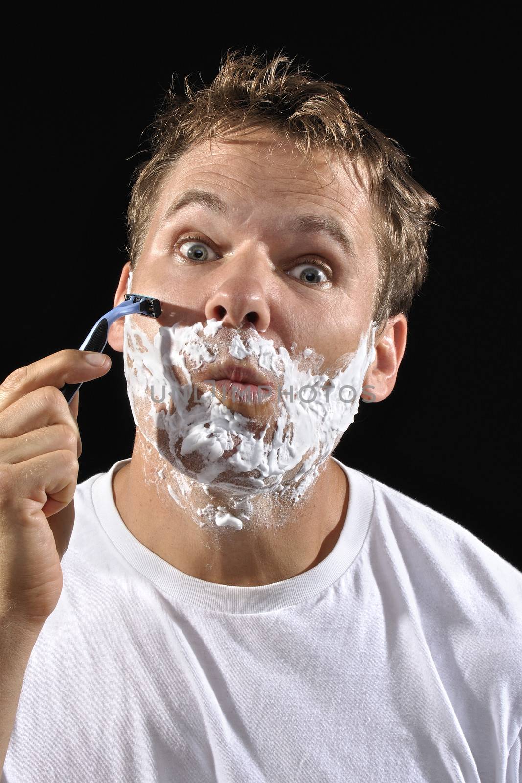 Handsome Caucasian man with messy bed head hair makes funny face while shaving his cheek on black background