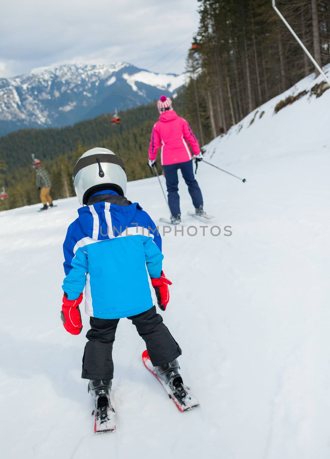 Happy little boy studying to ski in ski goggles and a helmet with his mother