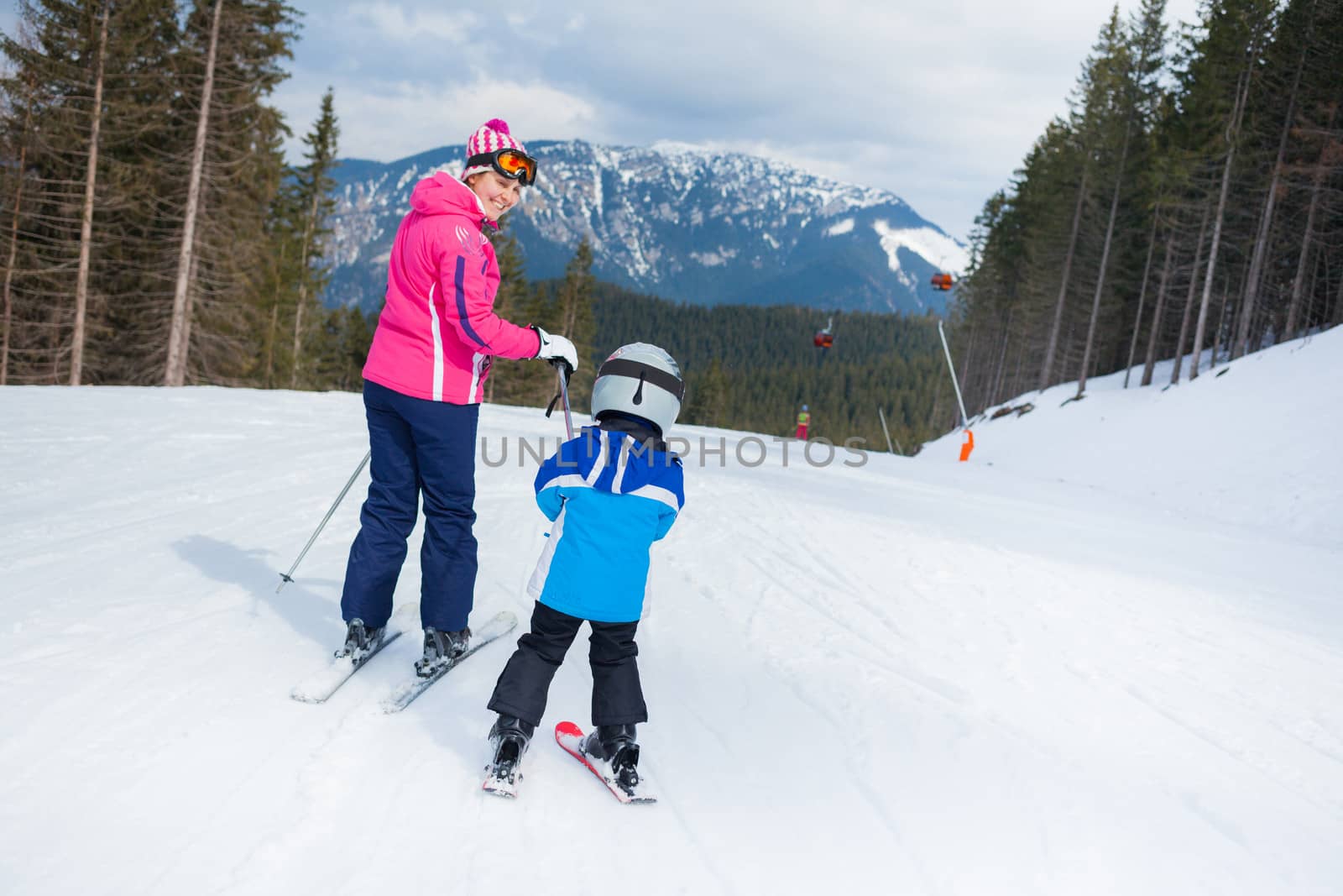 Happy little boy studying to ski in ski goggles and a helmet with his mother