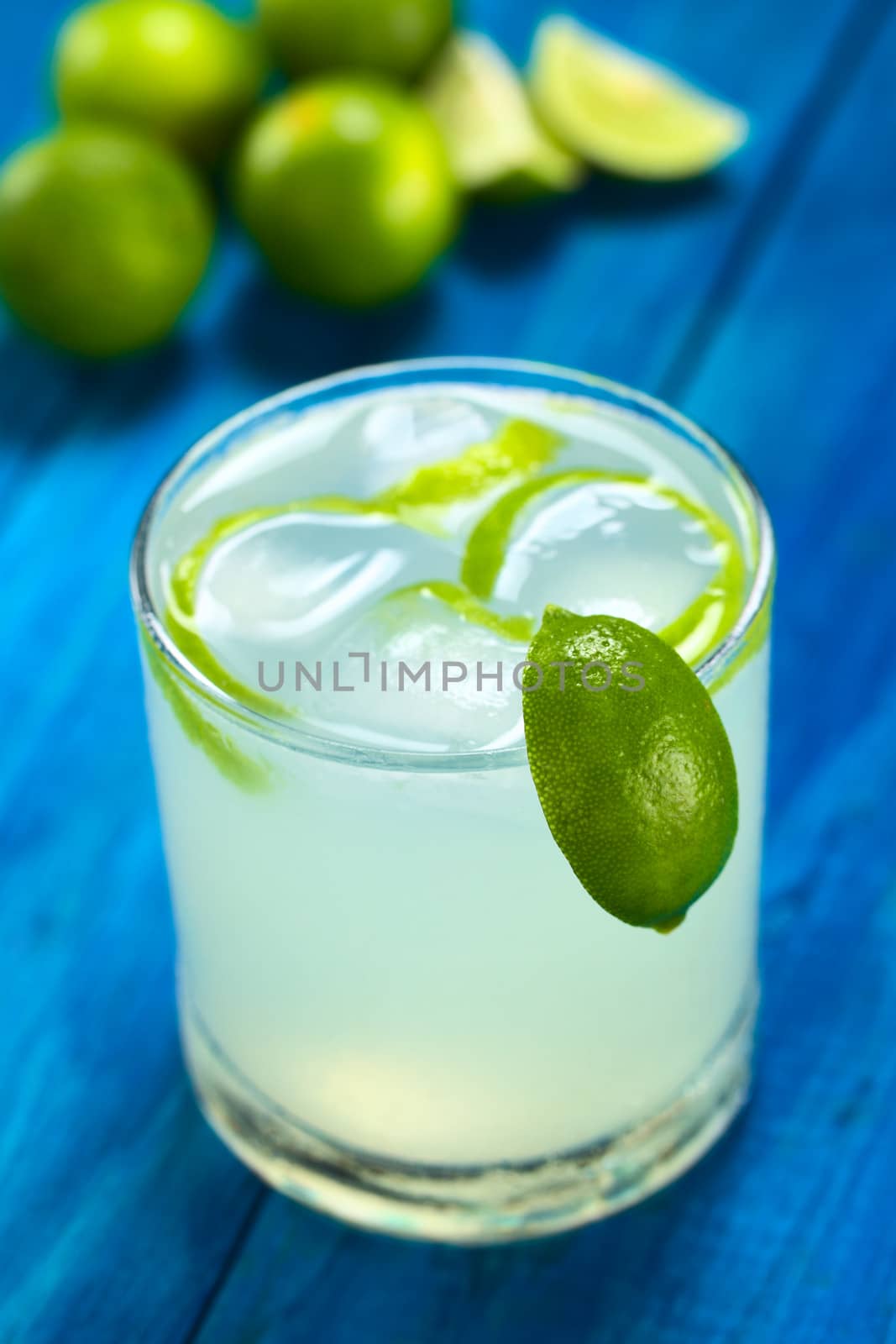 Freshly prepared refreshing lemonade out of limes with ice cubes and lime zest in glass garnished with a lime wedge on the rim; with limes in the back on blue wood (Selective Focus, Focus on the lime wedge on the rim)