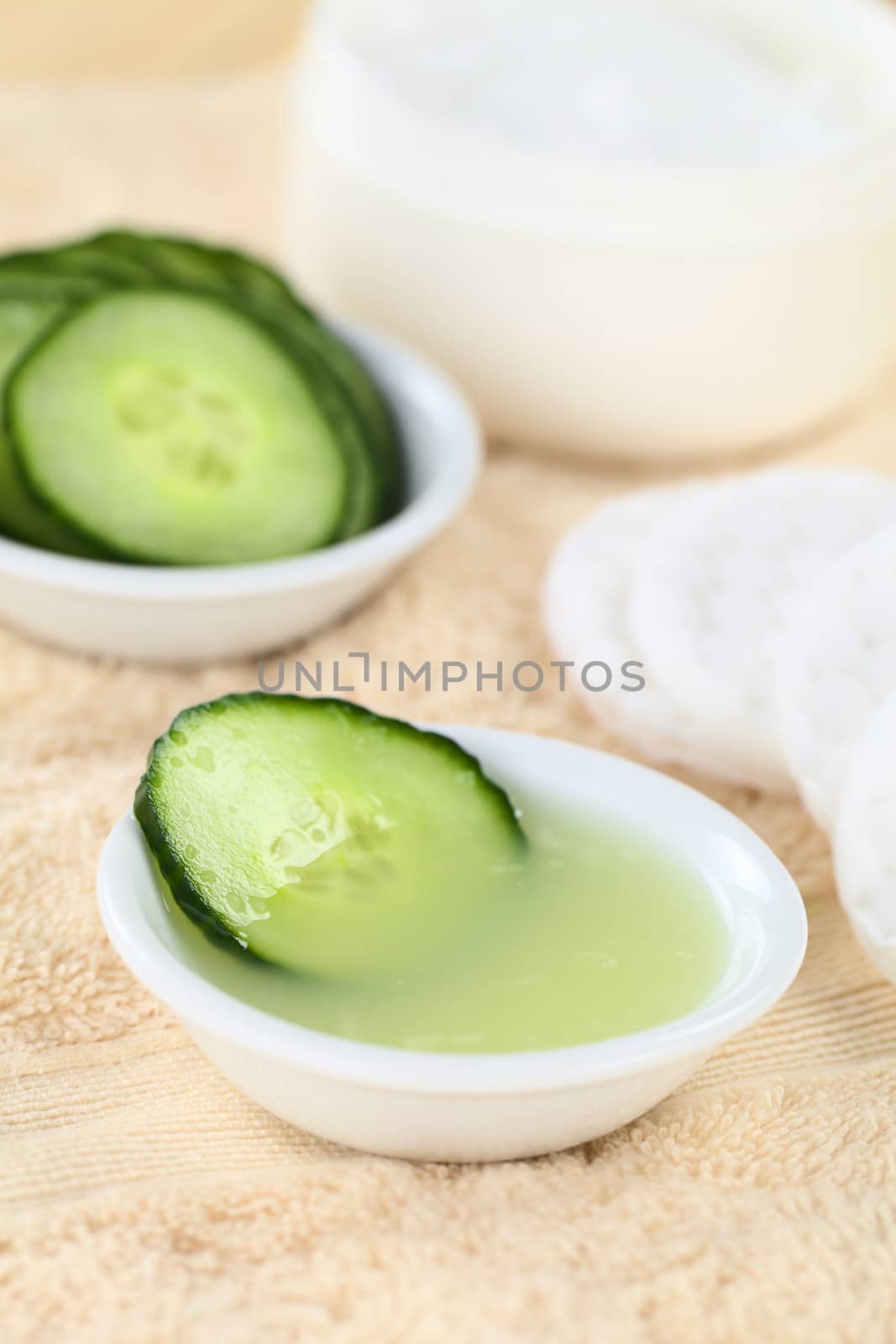 Homemade natural cucumber facial toner that hydrates, softens, soothes and cleanses the skin in a bowl with a cucumber slice, with facial pads and cream on towel (Selective Focus, Focus on the front of the cucumber slice in the toner)