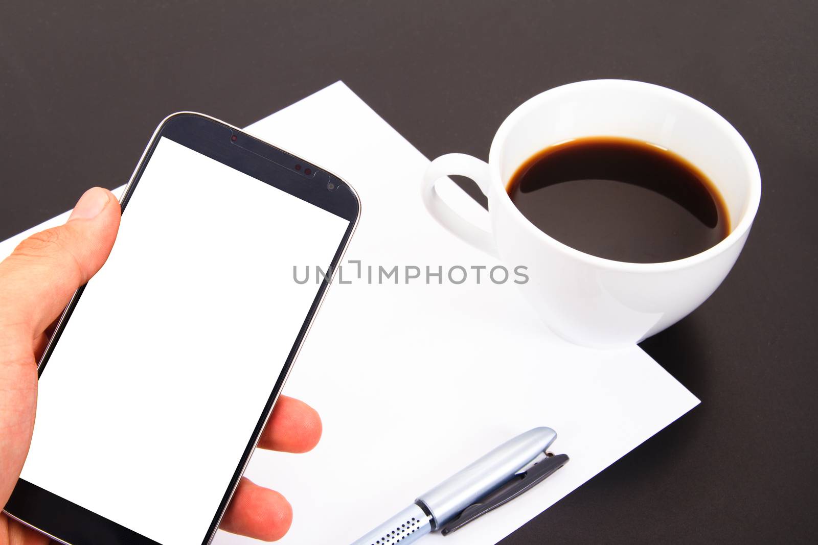 Hand showing smart phone with white, blank screen and paper, pen, coffee cup on wooden table.