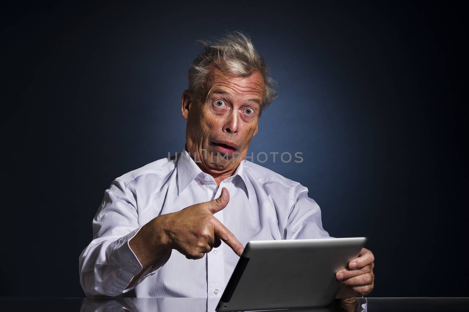 Shocked senior man pointing to his tablet computer with his finger with a look of appalled disbelief and confusion, humorous upper body studio portrait