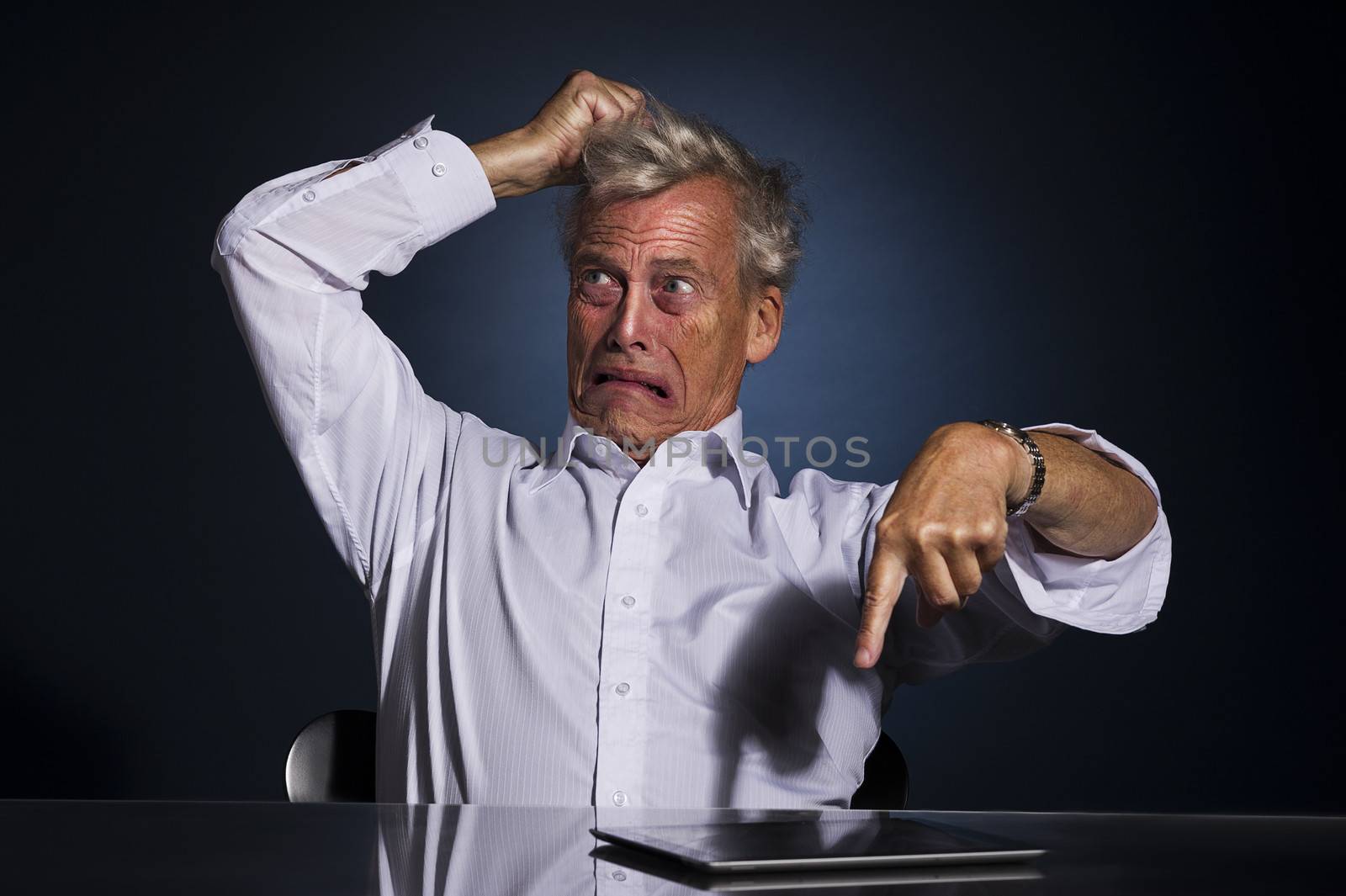 Very emotional upset frustrated senior business man pointing at his tablet computer lying on the desk while tearing at his hair with his hand