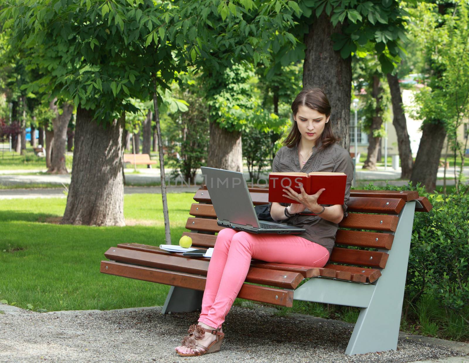 Young woman with a laptop reading a book and sitting on a bench outside in a park in summer.