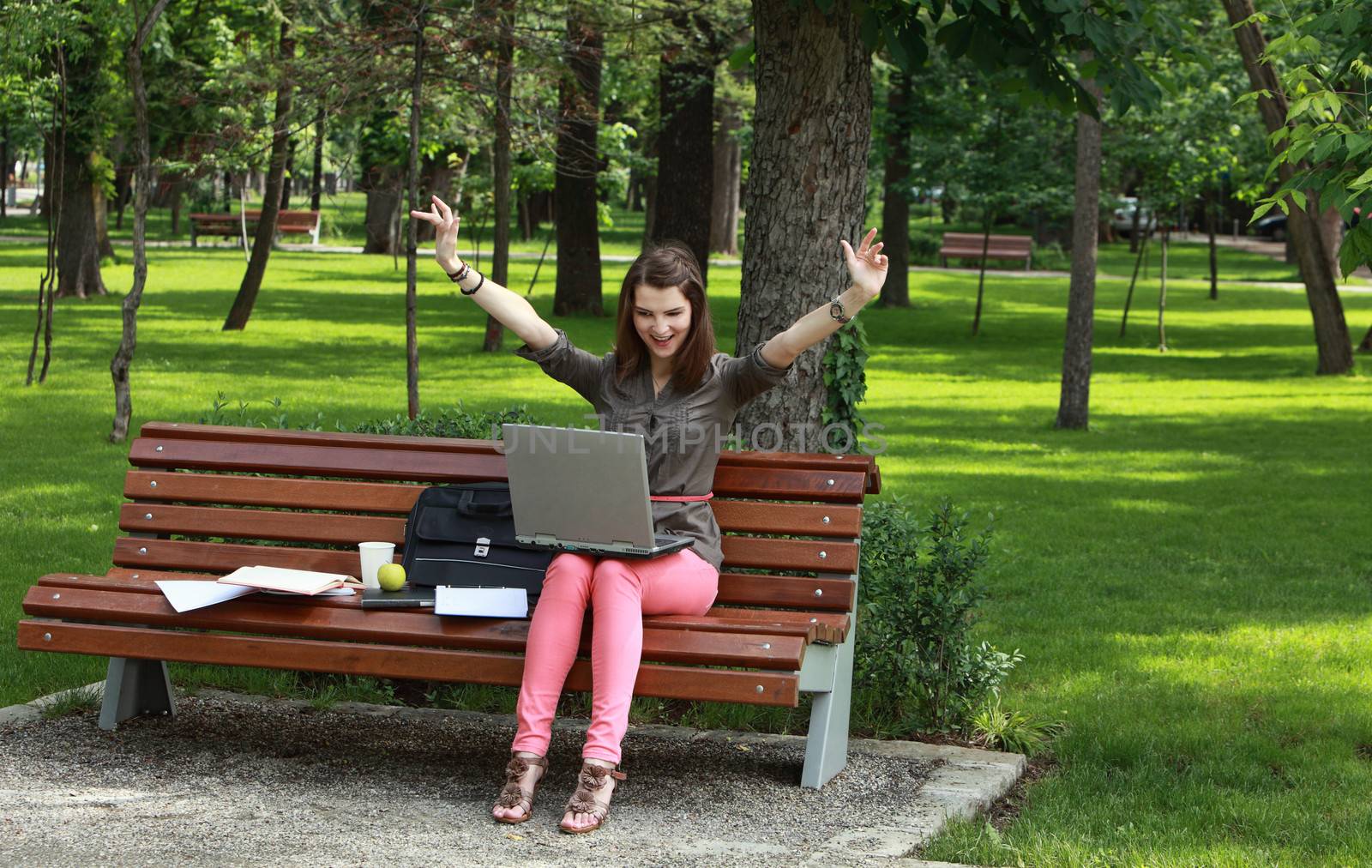 Happy young woman with a laptop rising her arms up, outside in an urban park.