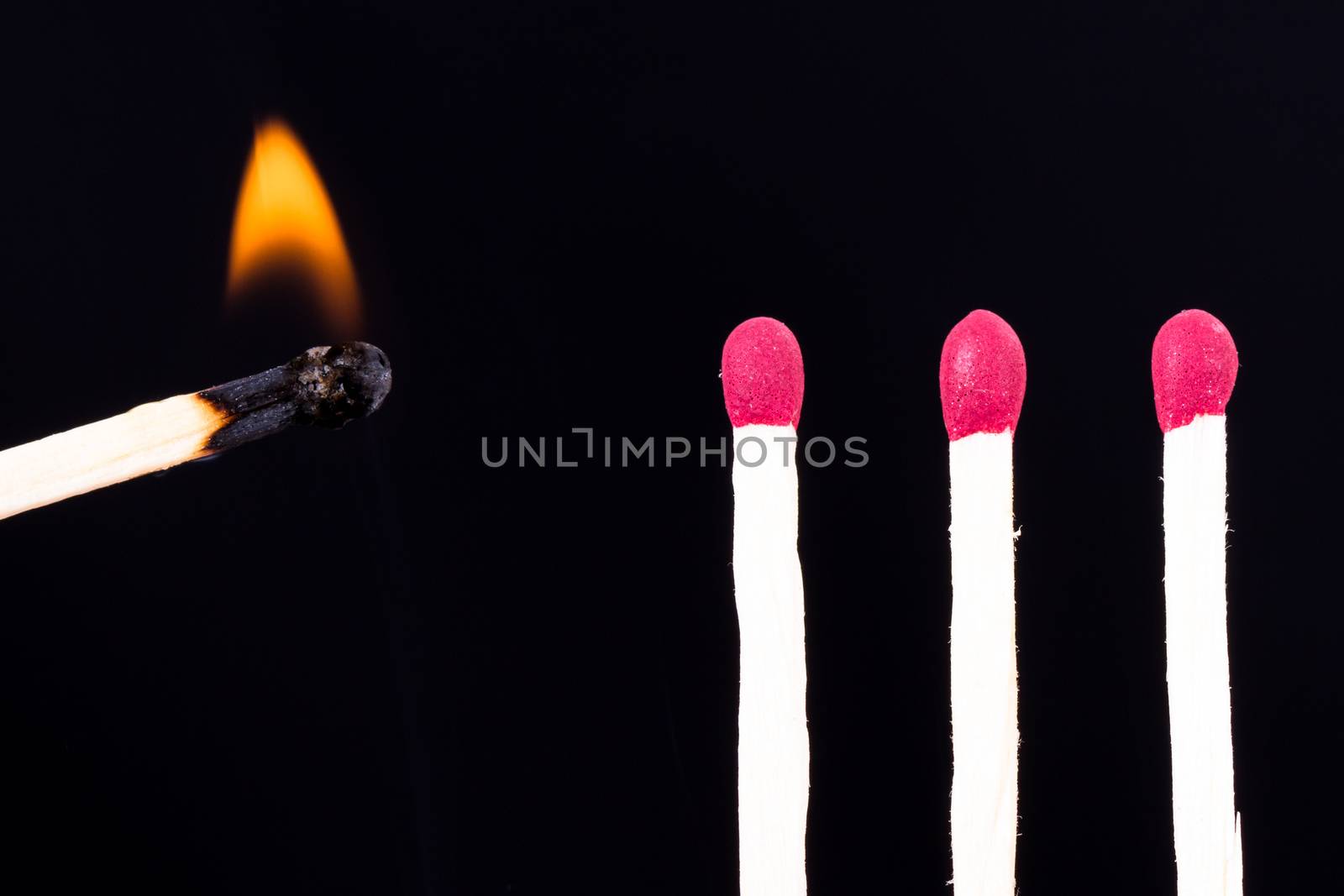 Burning match approaching red headed matches on dark background.