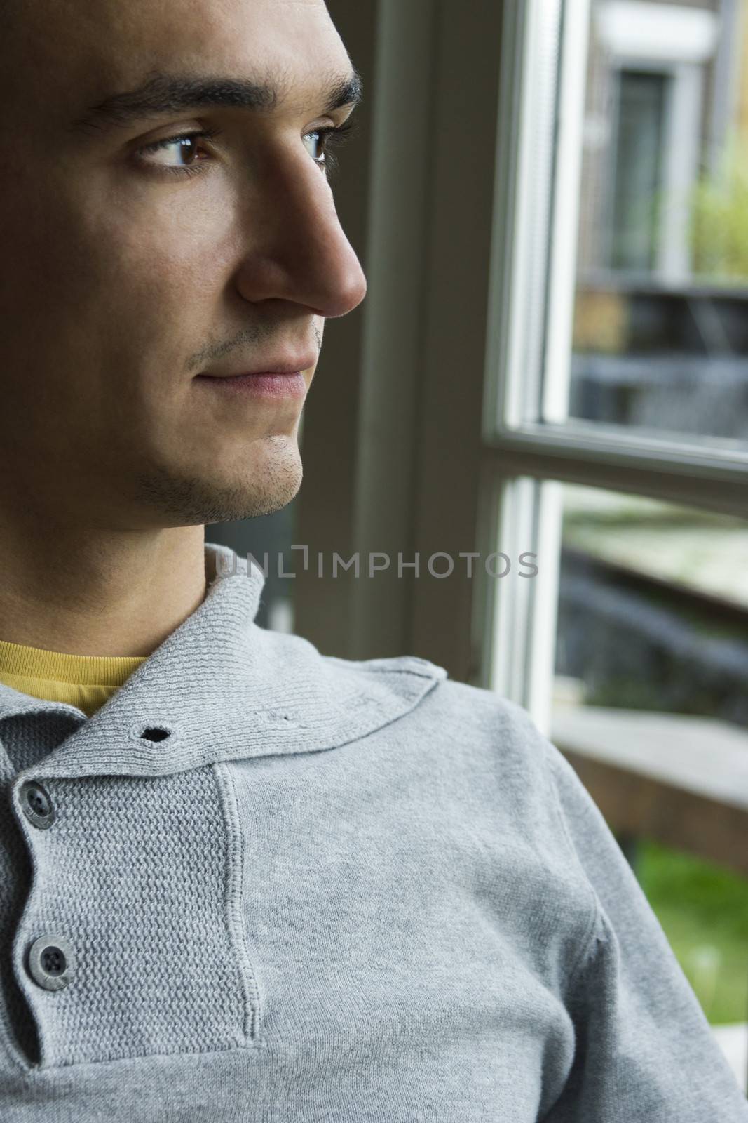 Serious pensive young man sitting in profile near the window, close-up face.