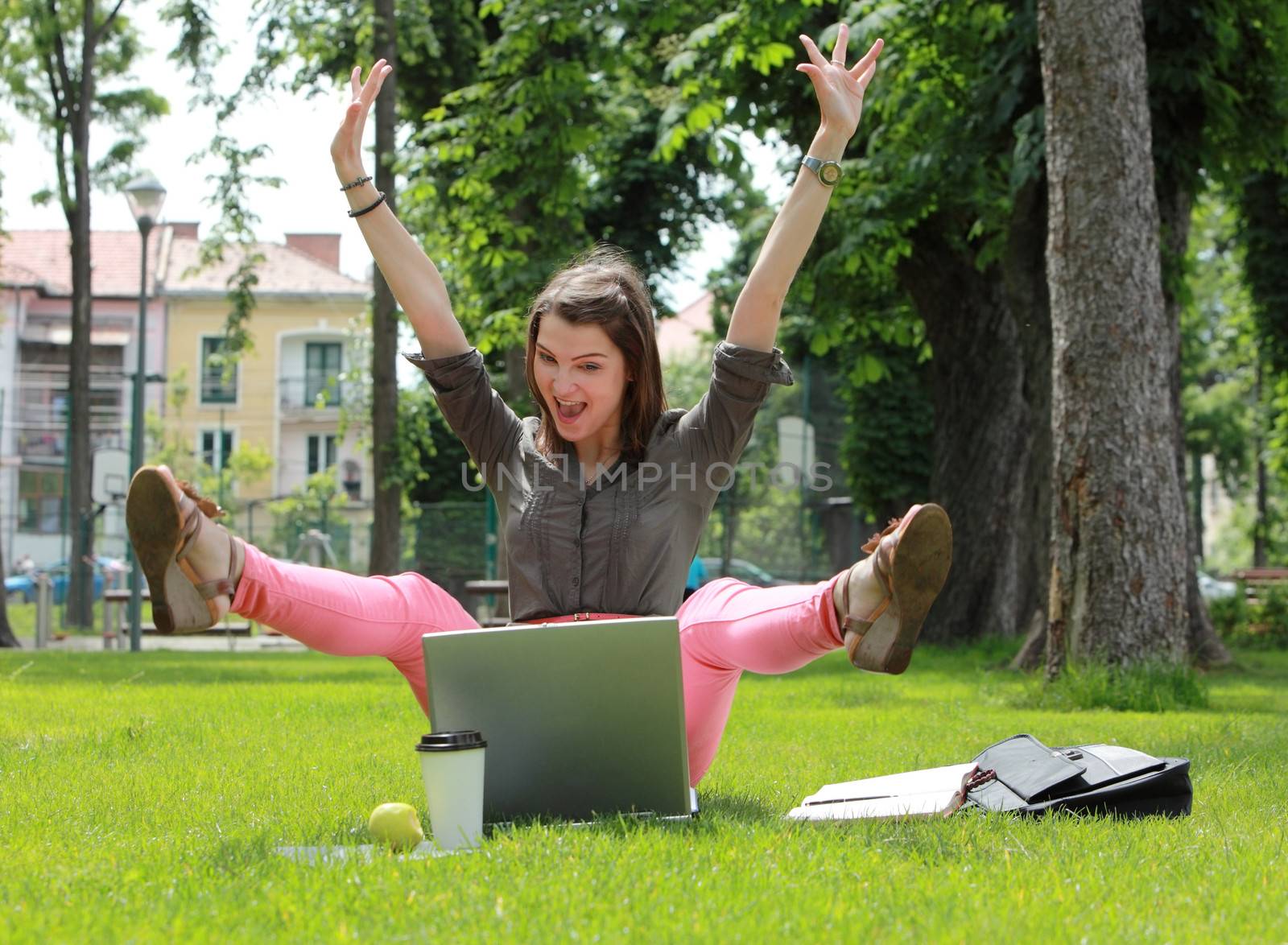 Excited happy woman with a laptop raising her hands and legs up in an urban park.