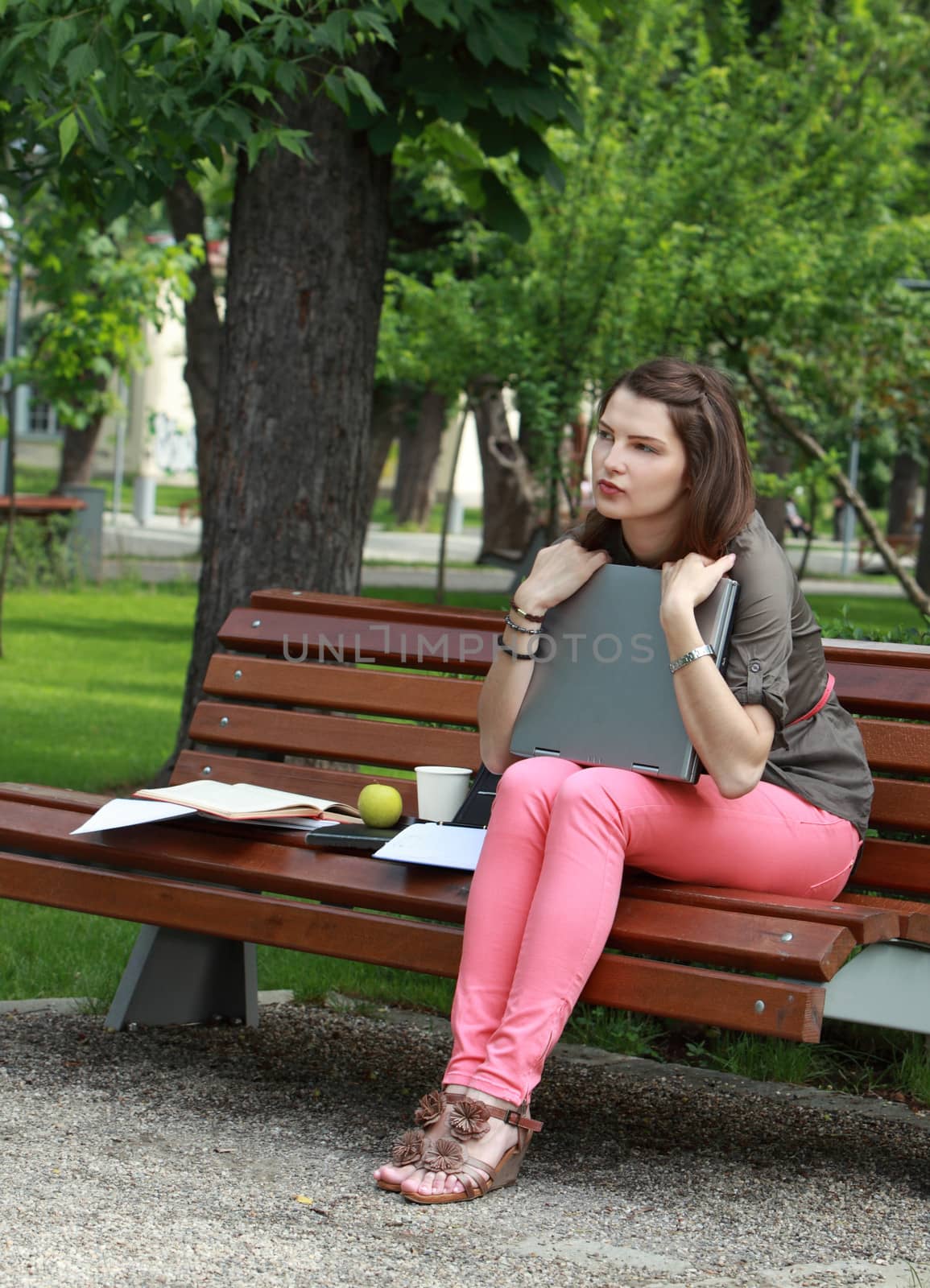 Young student woman hugging her laptop while sitting on a bench in a park.