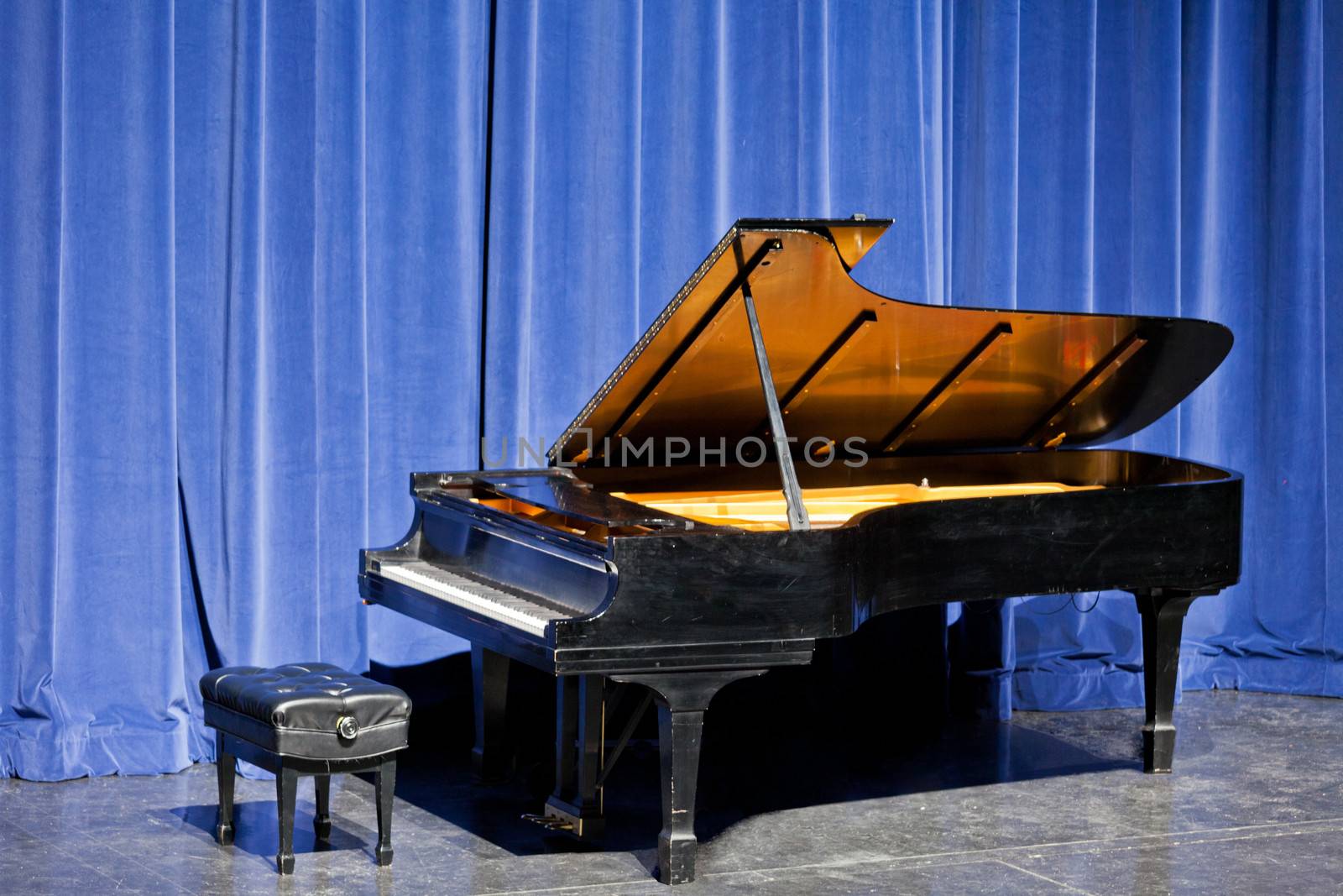 Open ebonised grand piano and piano stool standing in front of blue velvet curtains on stage ready for a musical recital or performance