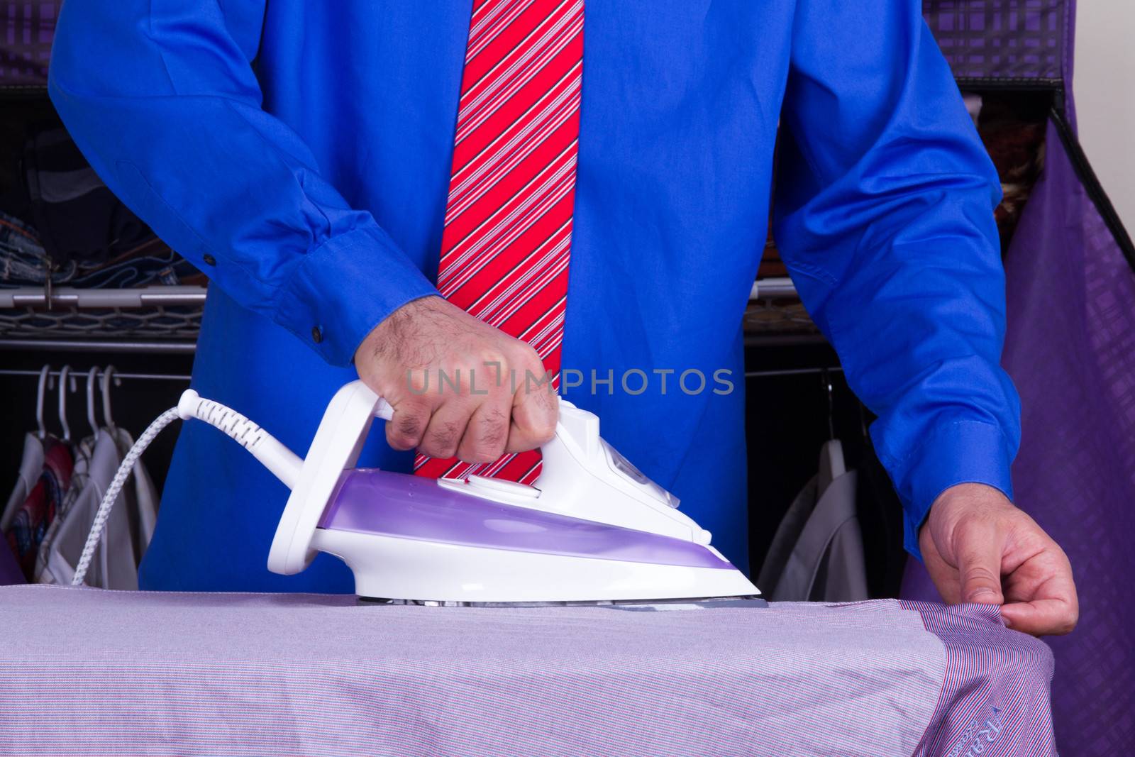Businessman with red tie and blue shirt ironing shirt against wardrobe.
