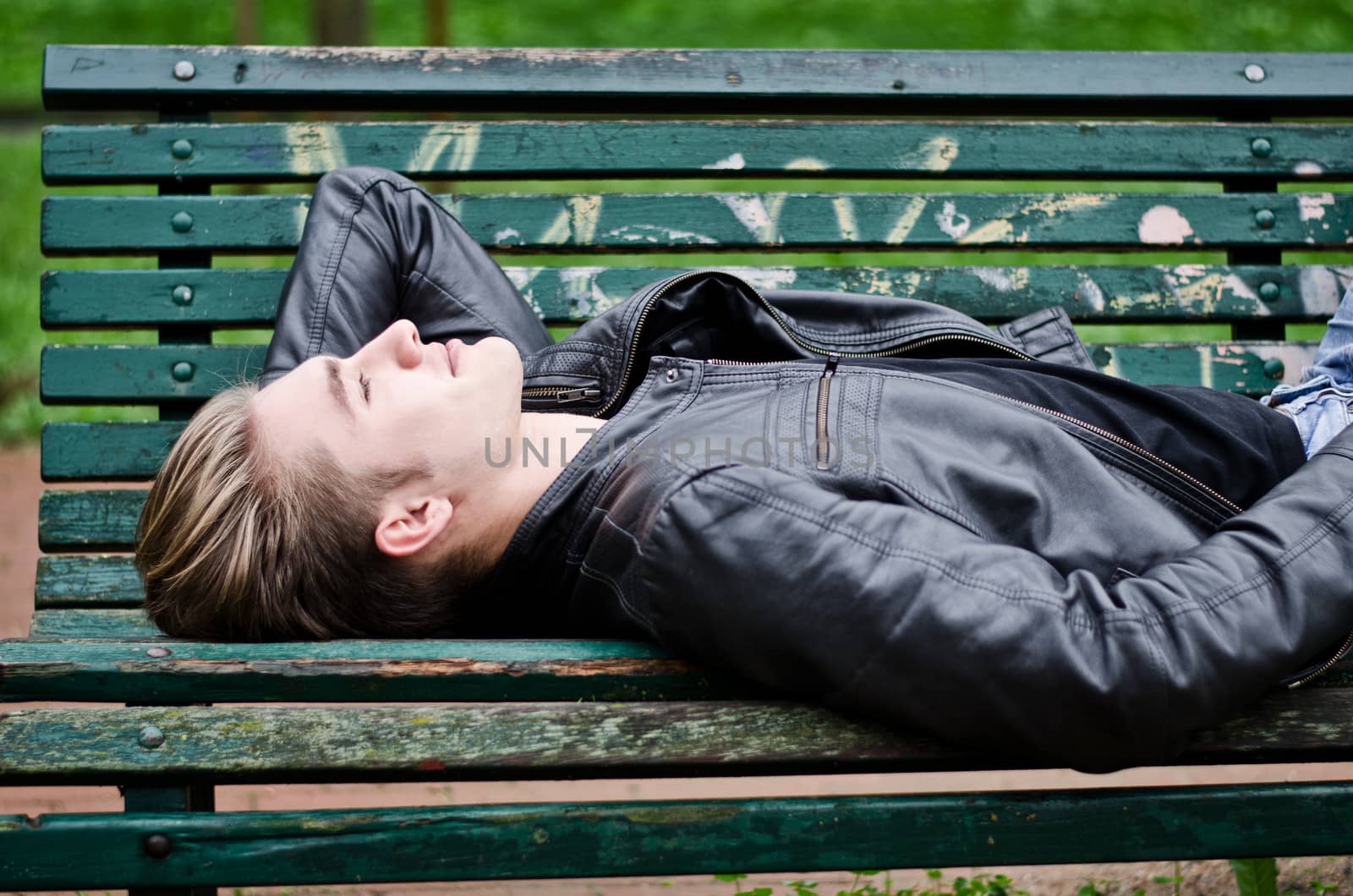 Handsome blond young man lying down on green, wooden park bench, looking up