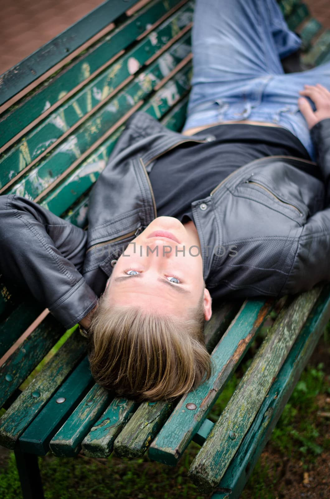 Handsome blond young man lying down on green, wooden park bench, looking at camera