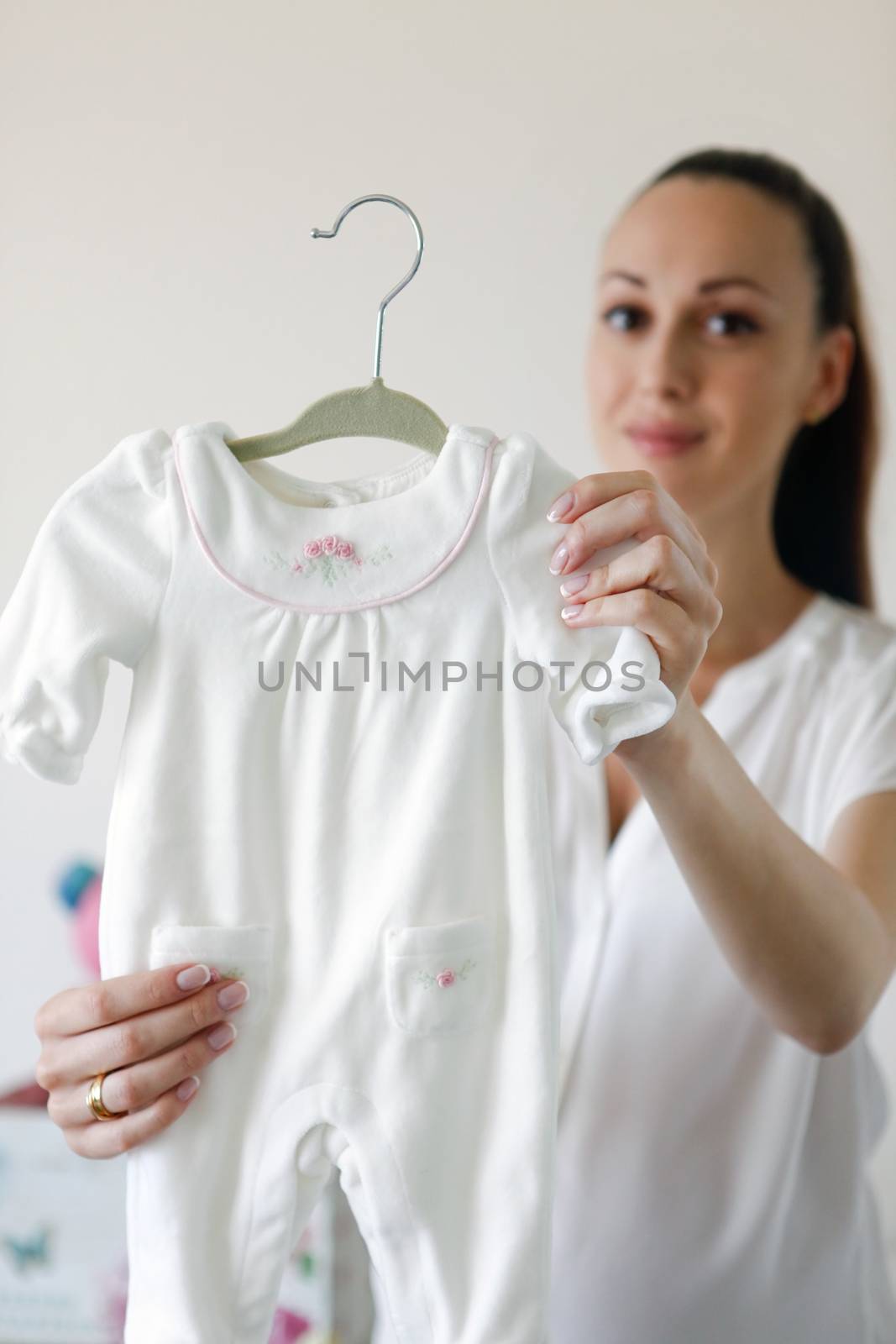 Happy beautiful pregnant woman with baby clothes in hands at home