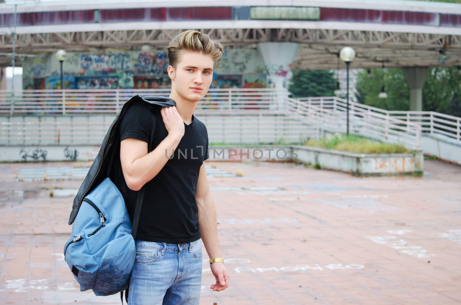 Handsome blond, blue eyed young man outdoors with ruck sack on shoulder, t-shirt and jeans