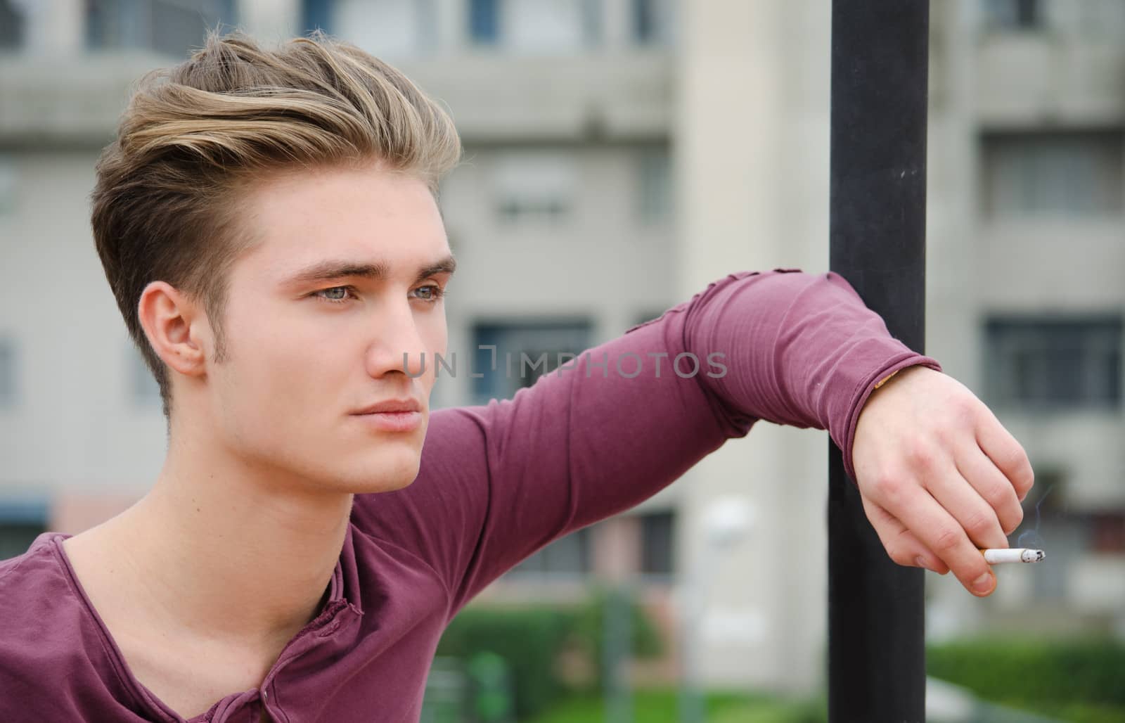 Attractive blue eyed, blond young man smoking cigarette outdoors leaning against pole