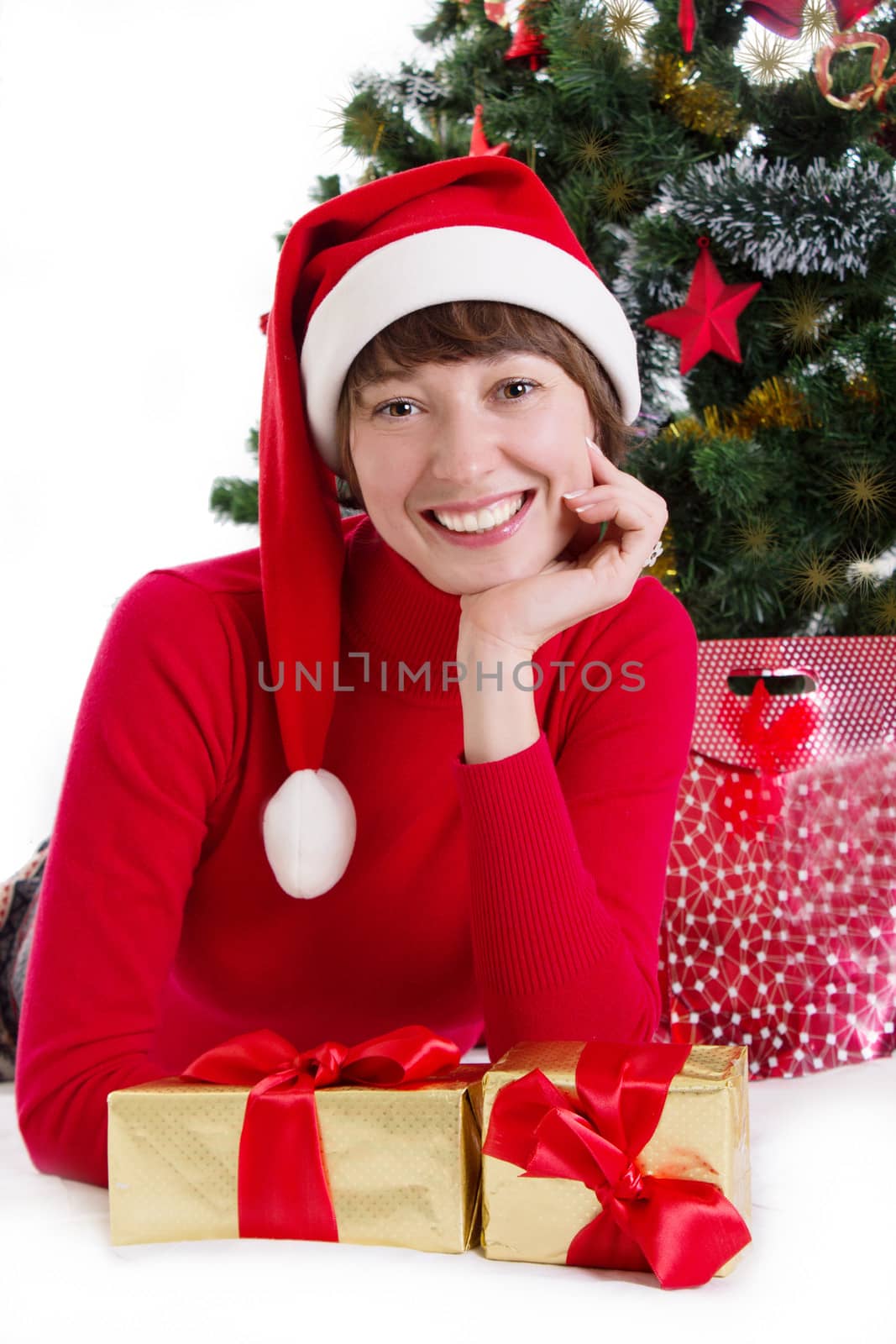 Smiling woman in red Santa hat lying under Christmas tree with gifts